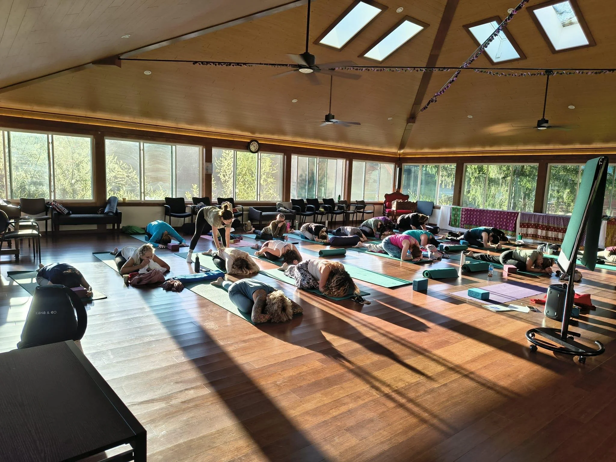 Yoga class in a bright, spacious room with large windows, ceiling fans, and skylights. Participants are practicing yoga on mats, led by an instructor. The room has a wooden floor and a clock on the wall.