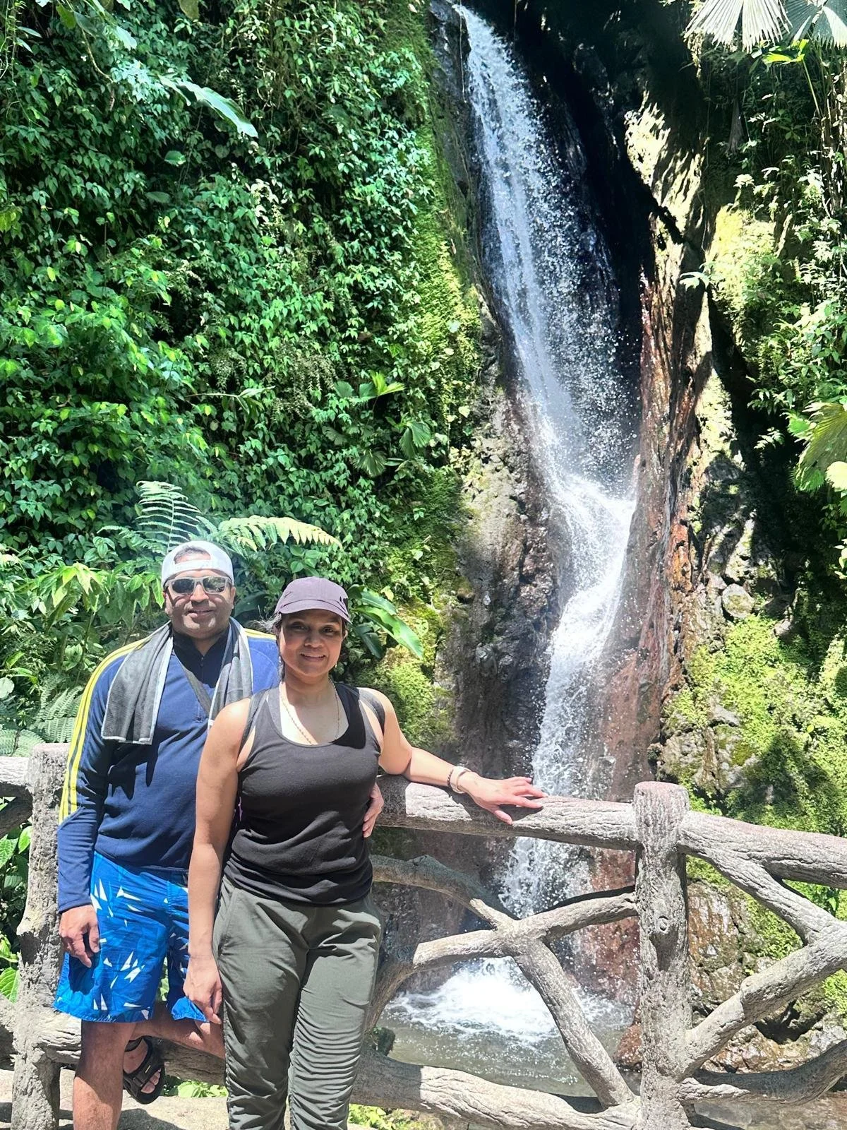 A man and woman standing near a wooden railing in front of a waterfall surrounded by green foliage.
