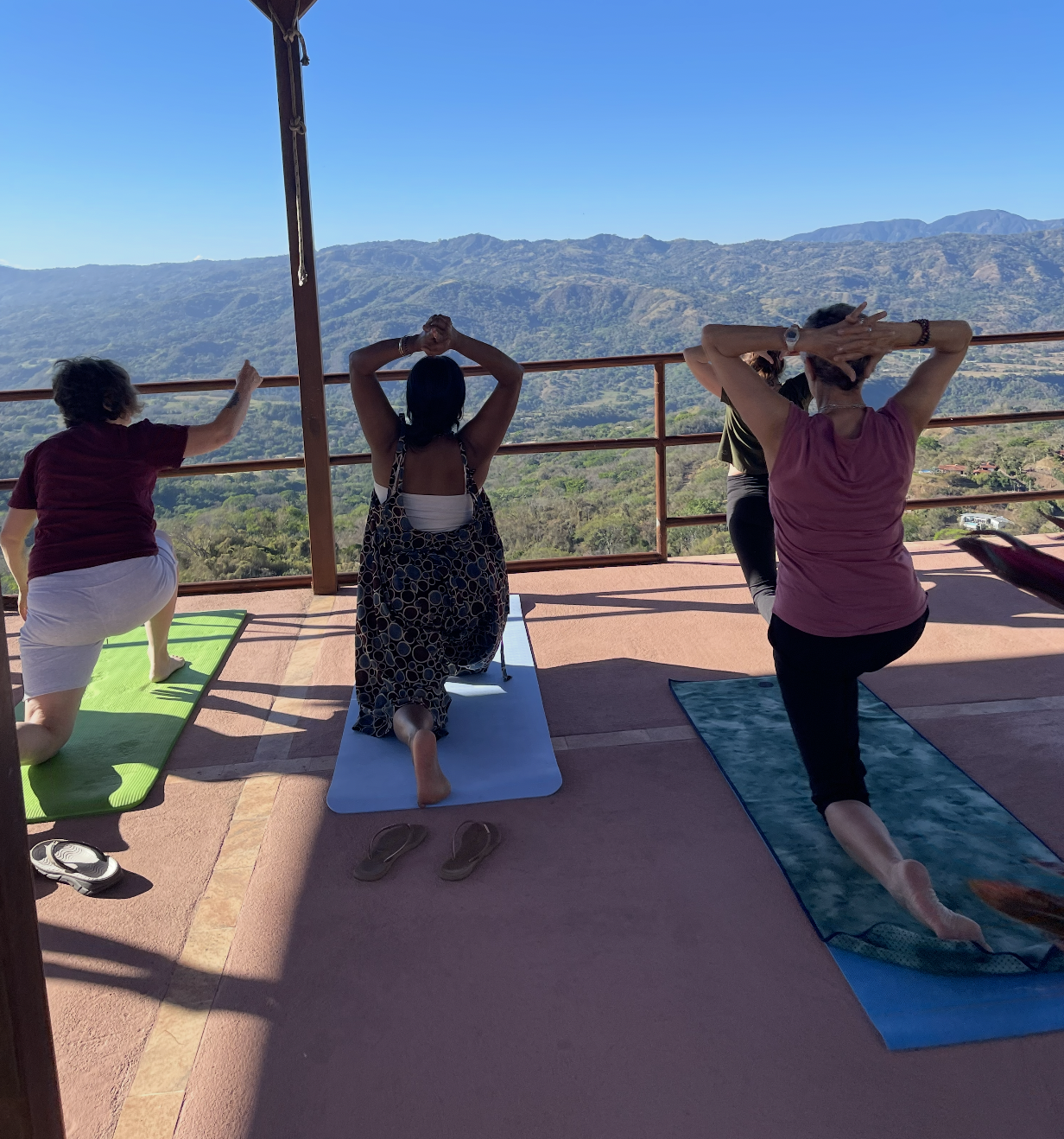 Four women practicing yoga on mats on a balcony with mountain view, some kneeling and stretching.