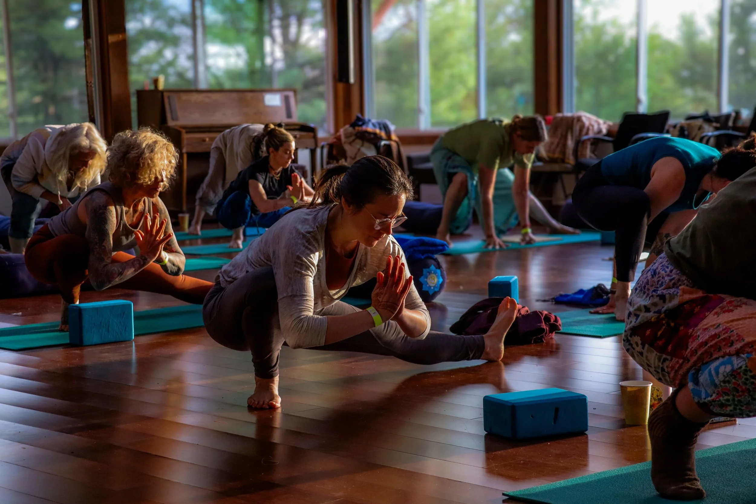 A group of people practicing yoga in a spacious room with large windows showing greenery outside, some are in a downward dog pose while others are in different positions, with yoga mats and blocks on the floor.