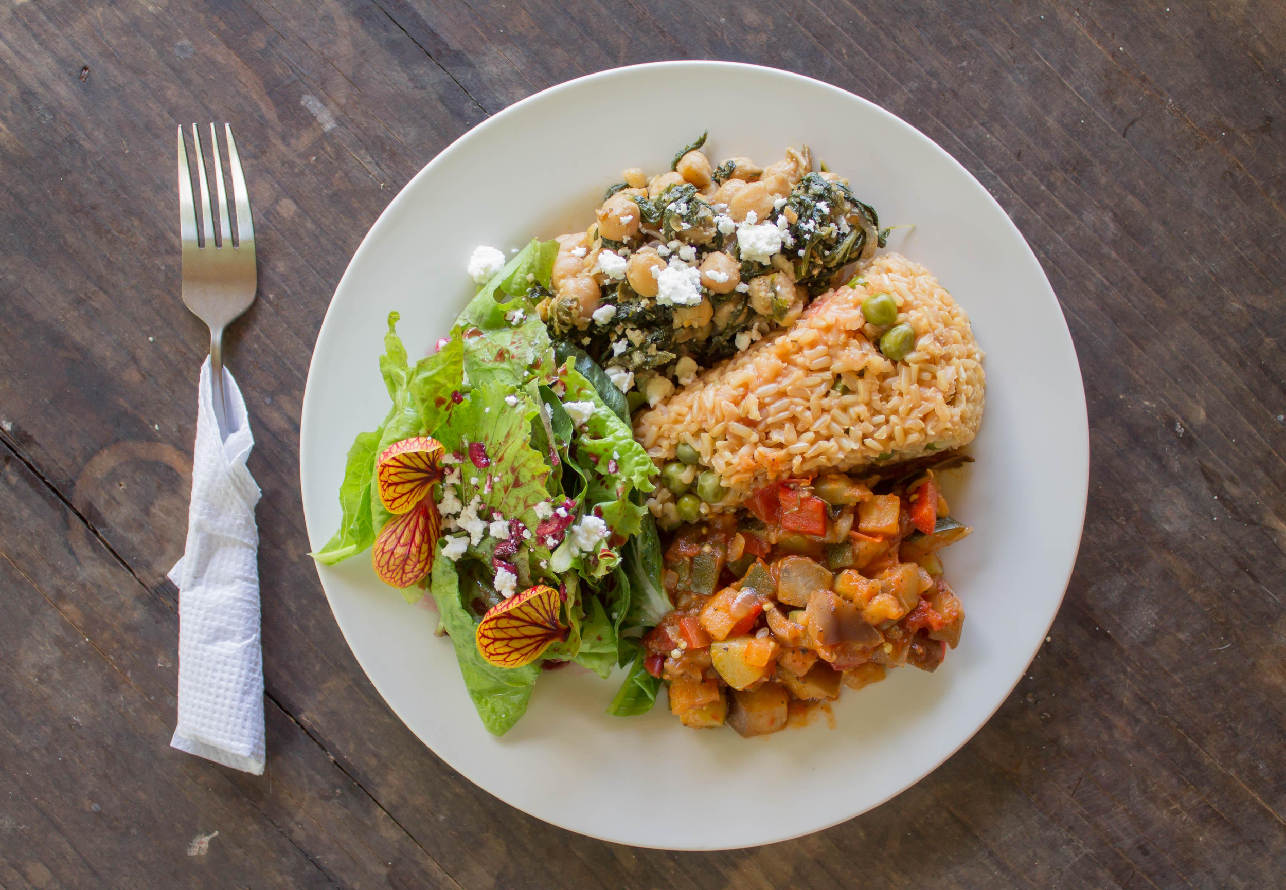A white plate with multiple servings of food, including a chickpea and vegetable stew, a portion of rice, and a small side salad with greens and edible flowers, on a dark wooden table, with a fork and a paper napkin to the left.