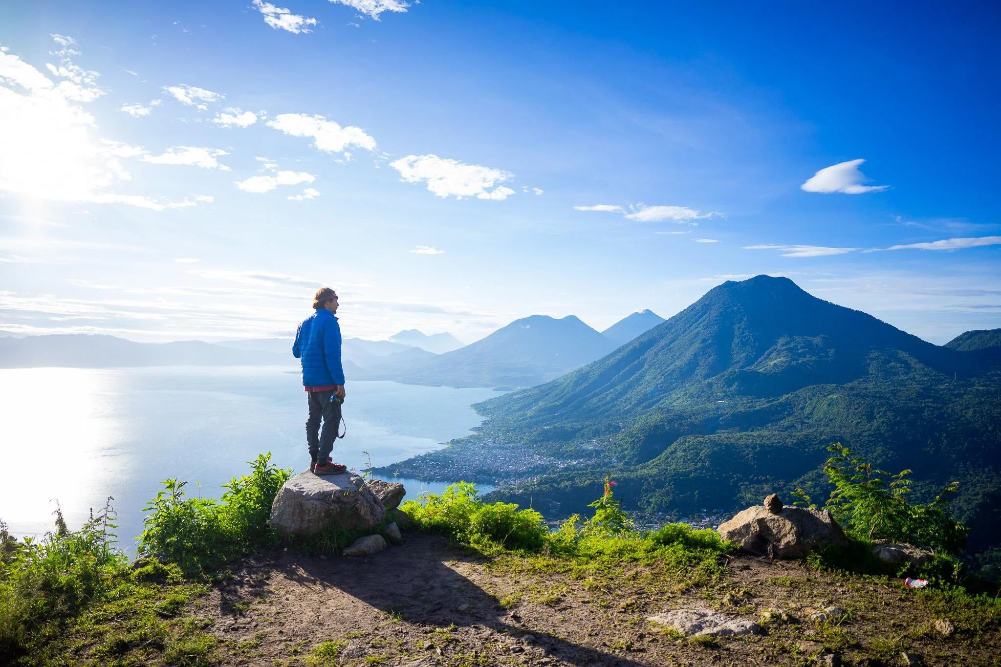 A person standing on a rocky ledge overlooking a large body of water and lush green mountains under a bright blue sky with scattered clouds.