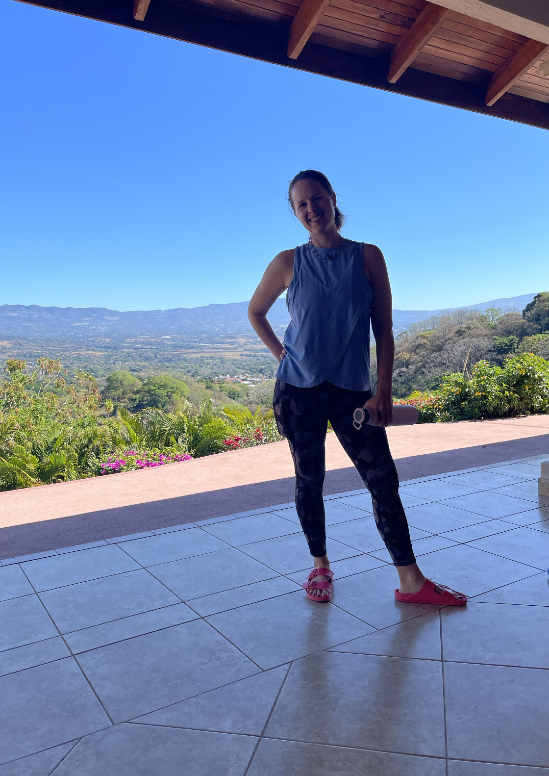 A woman smiling on a tiled porch with a scenic mountain view and bright blue sky in the background. She is wearing a sleeveless top, patterned leggings, and pink slides, holding a water bottle in her right hand.