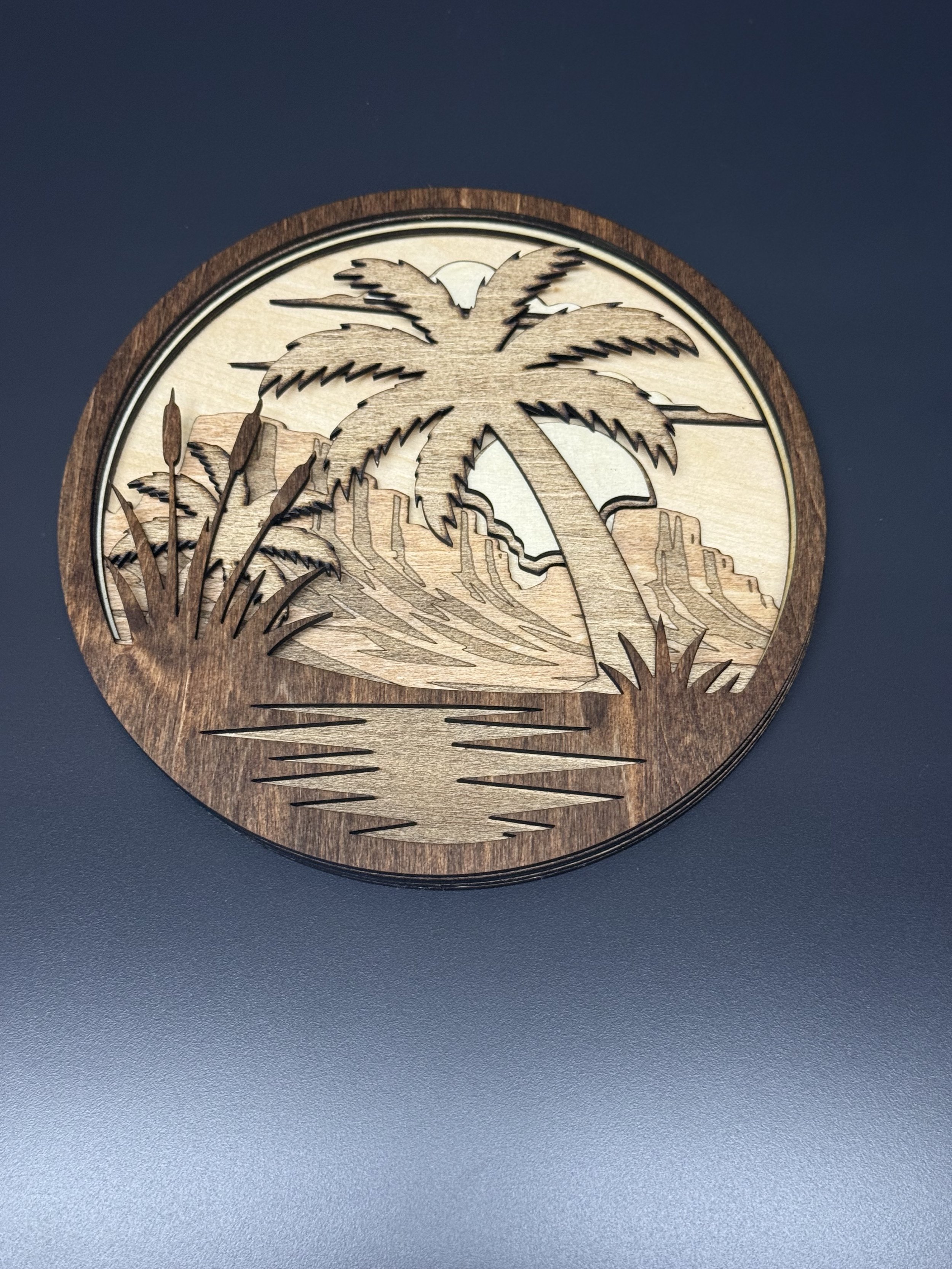 A wooden round wall hanging with a laser-cut design a desert oasis scene, including a palm tree, cattails.
