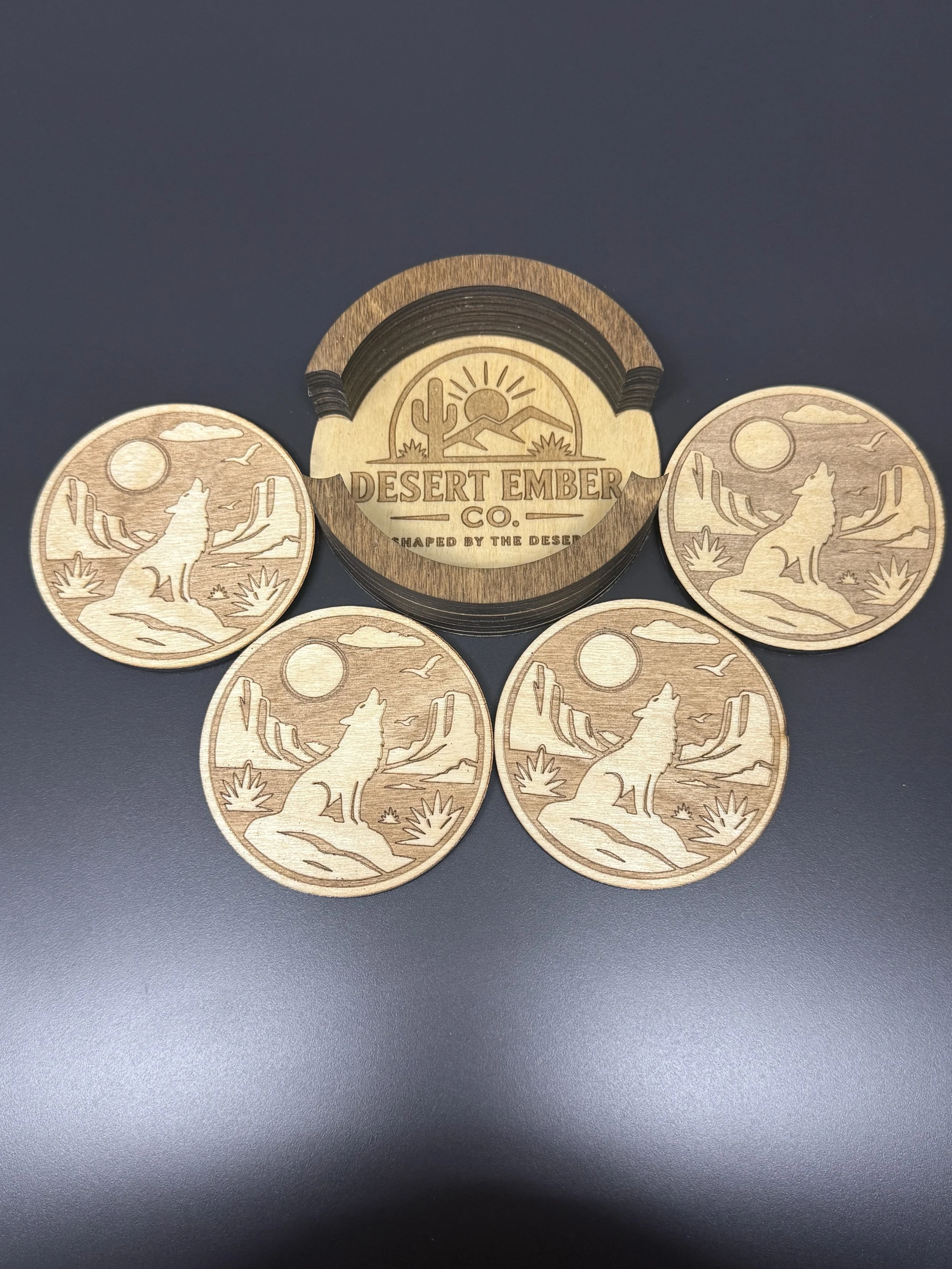 Wooden coasters with desert landscape design, including a howling coyote, mountains, cacti, and a sun, arranged on a dark surface.