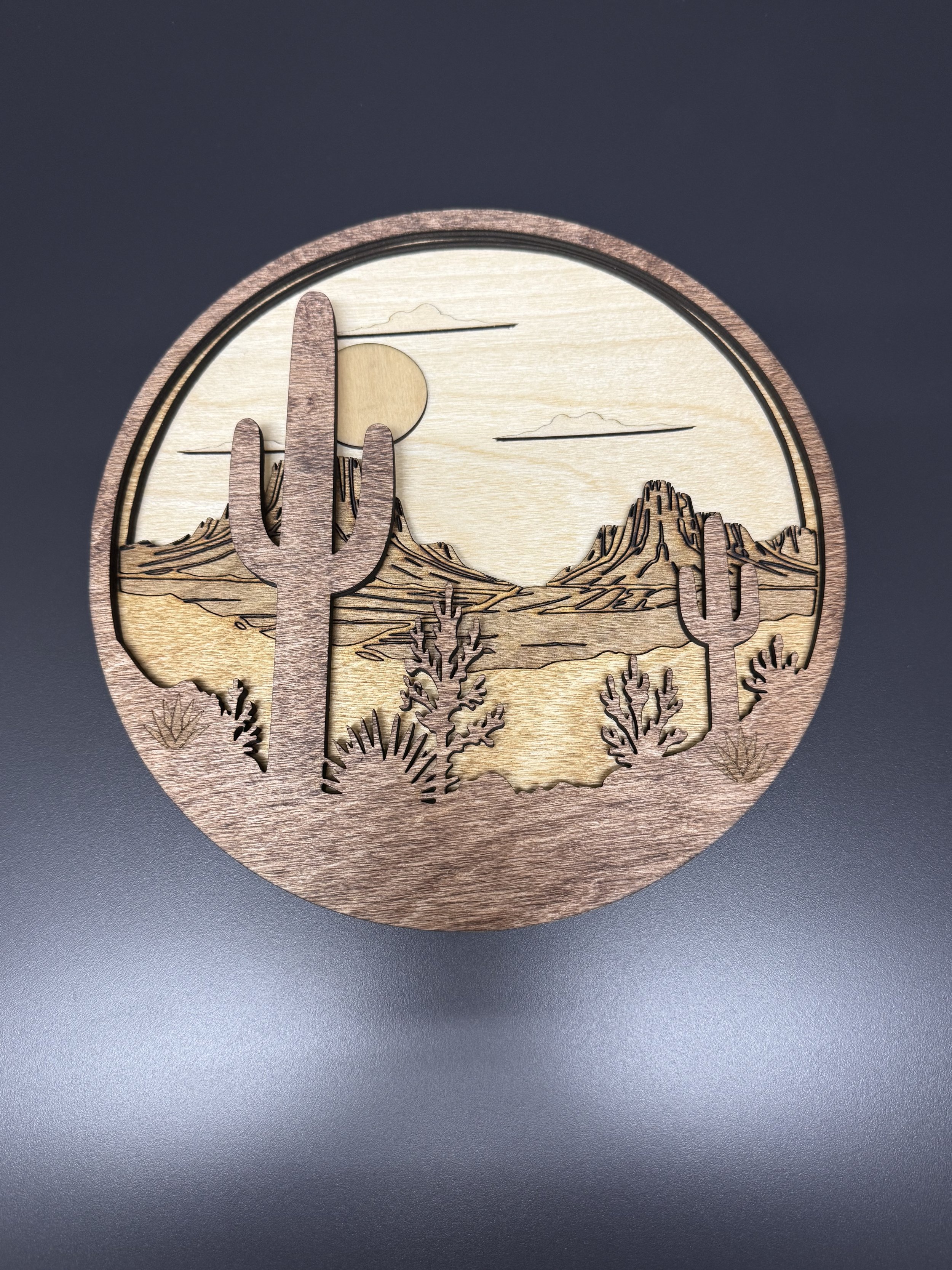 Wooden clock with desert landscape design, featuring cacti, mountains, a sun, and clouds, on a gray surface.