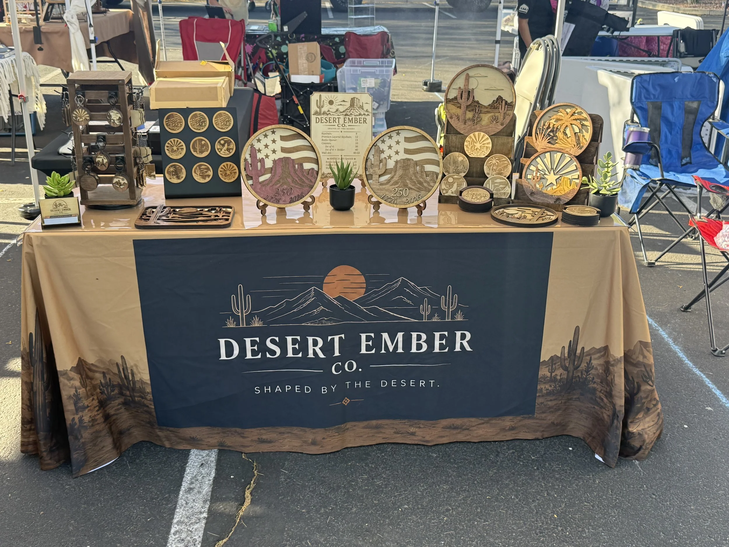 Outdoor market booth displaying wooden decorative items with desert landscape and cactus designs, a potted succulent, and a blue desert-themed banner.