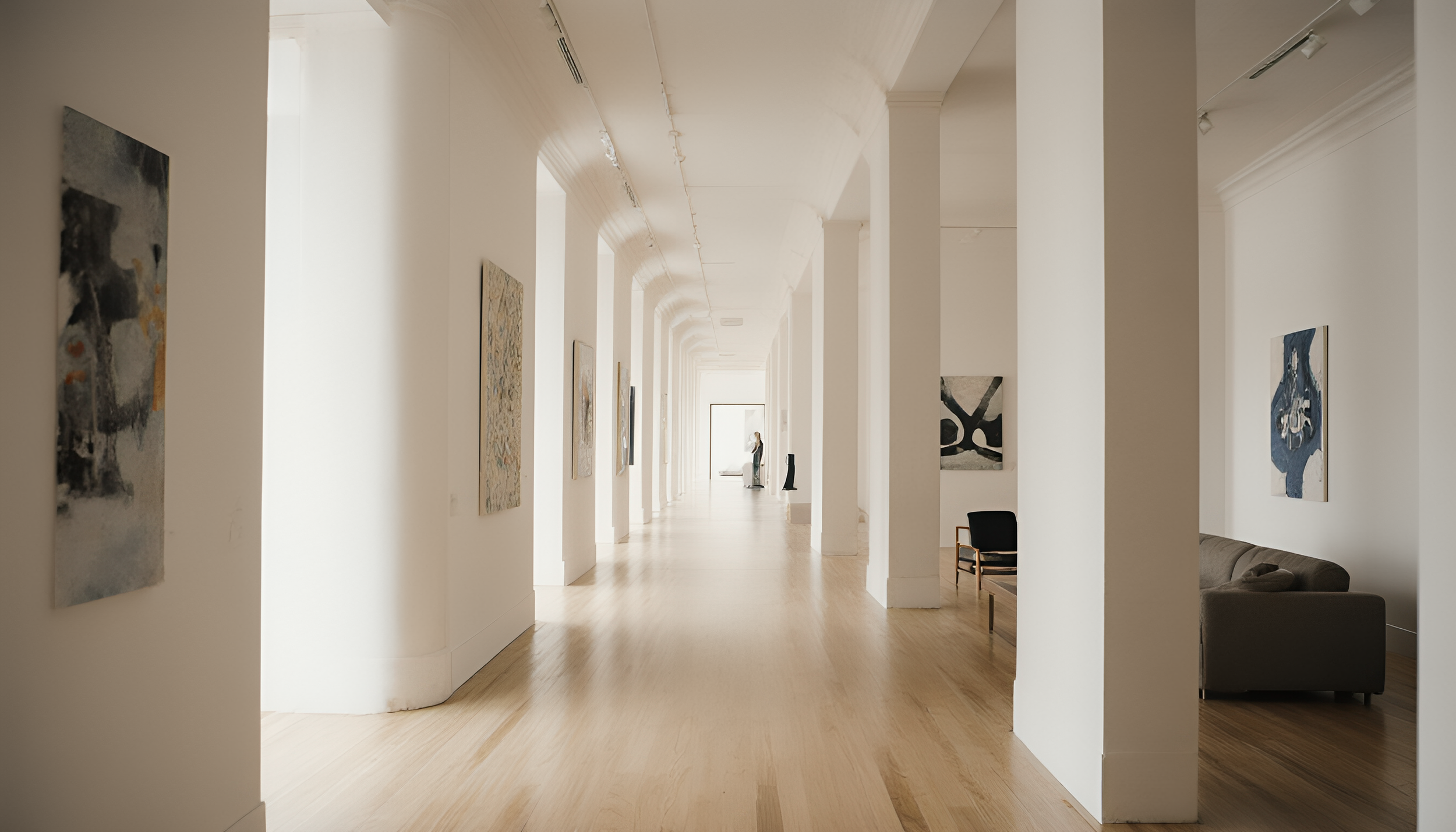 An art gallery with white walls and hardwood floors, featuring abstract paintings. A person is standing in the distance, observing the art. There is a sofa and chairs along the right side.