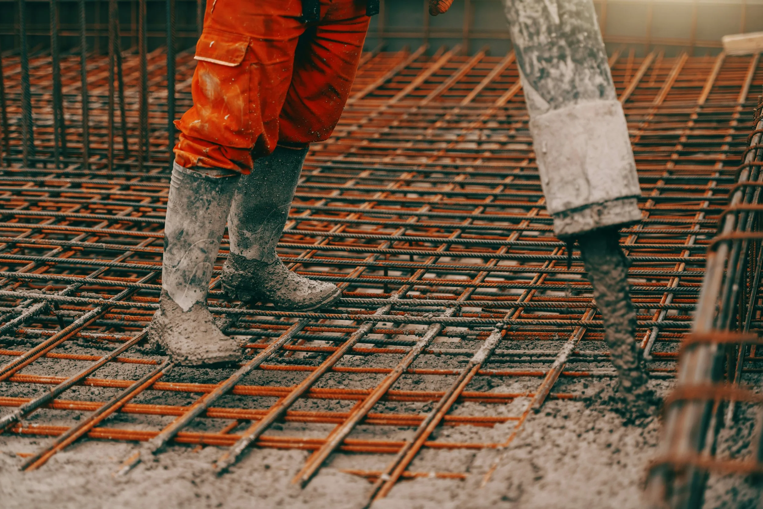 Close-up of a construction worker's legs and feet as they pour concrete over a rebar grid at a construction site.