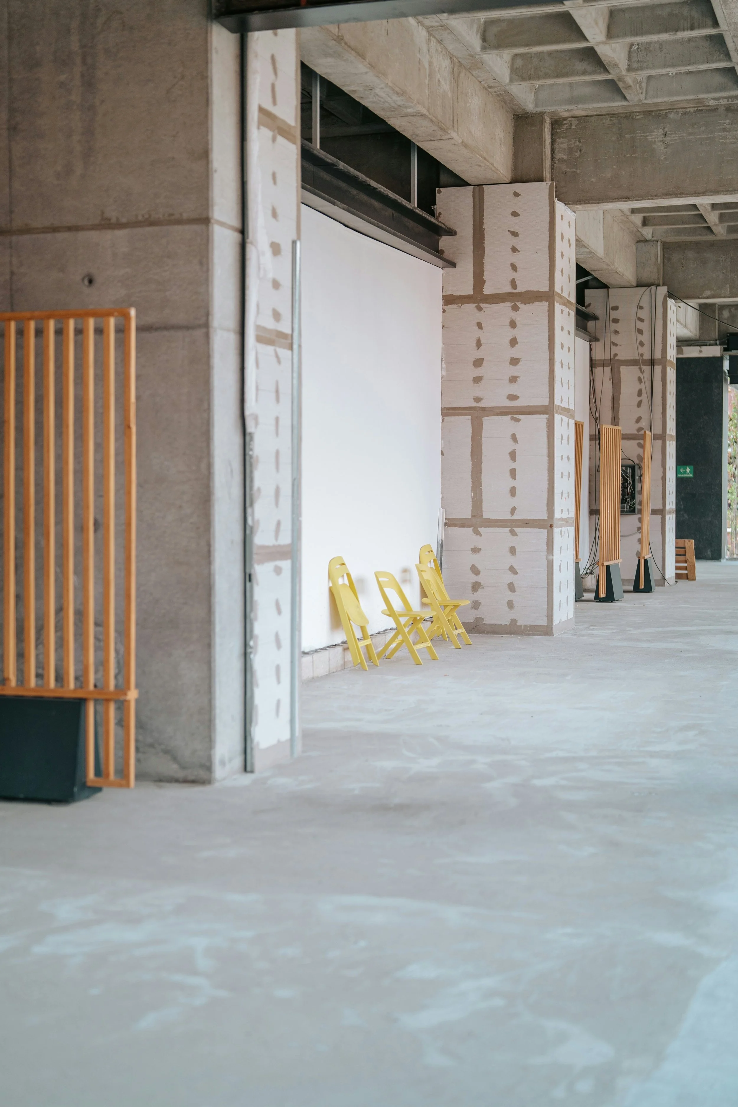 Interior view of a building under construction with bare concrete walls and floors, yellow plastic chairs lined along the wall, wooden slats, and construction materials and wires visible.