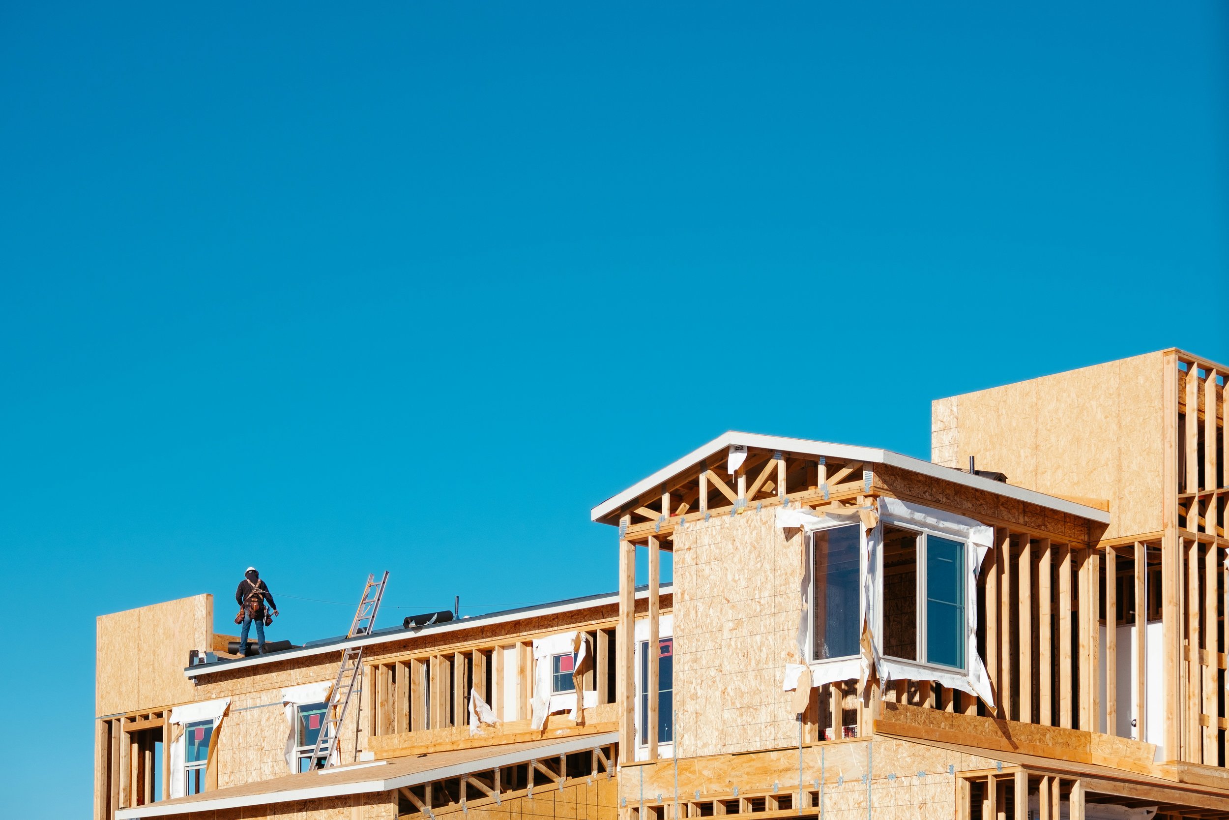 A house under construction with unfinished wooden framing and a worker standing on the roof, against a clear blue sky.