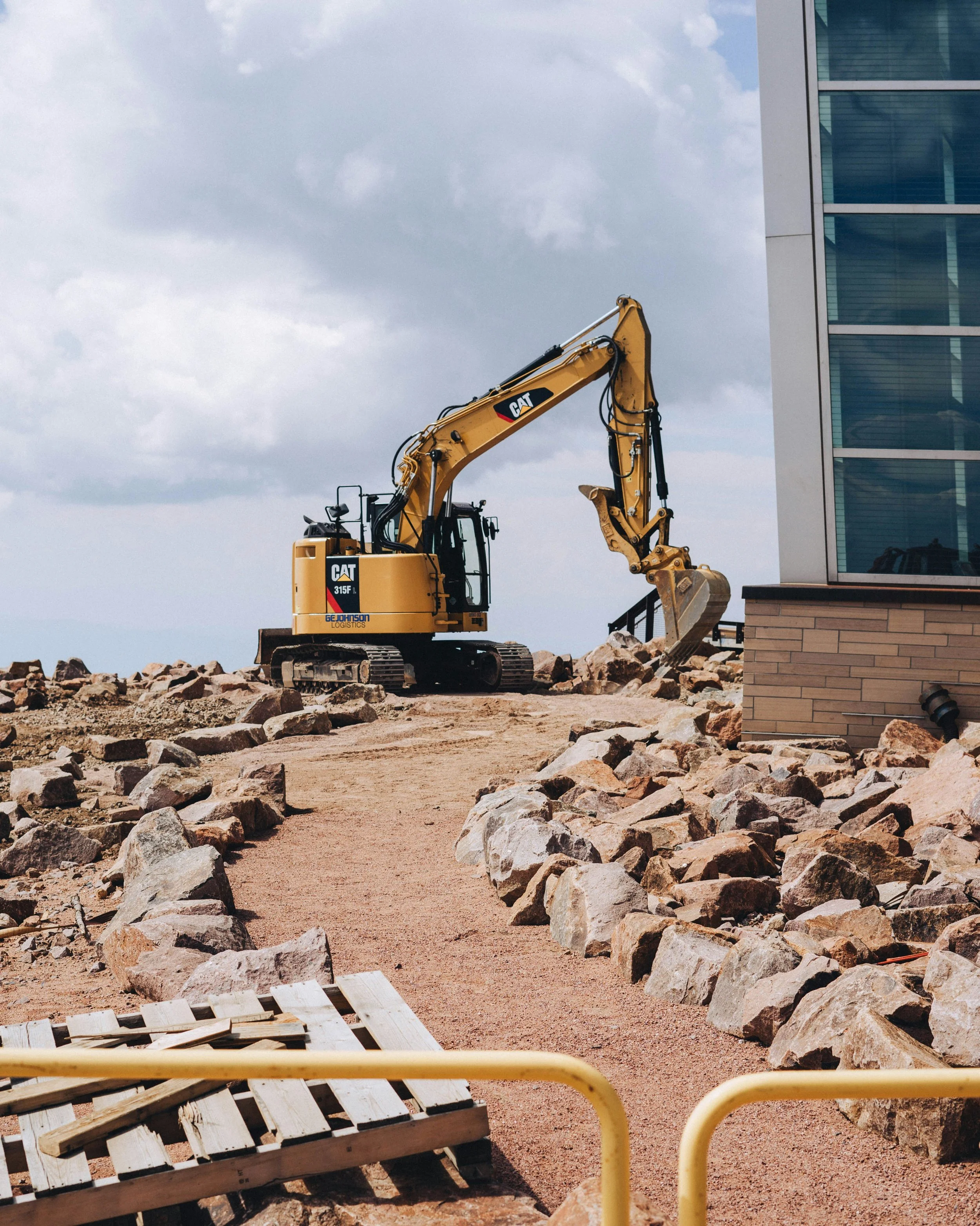 Construction site with a yellow CAT excavator working on a rocky terrain near a modern building with glass windows.