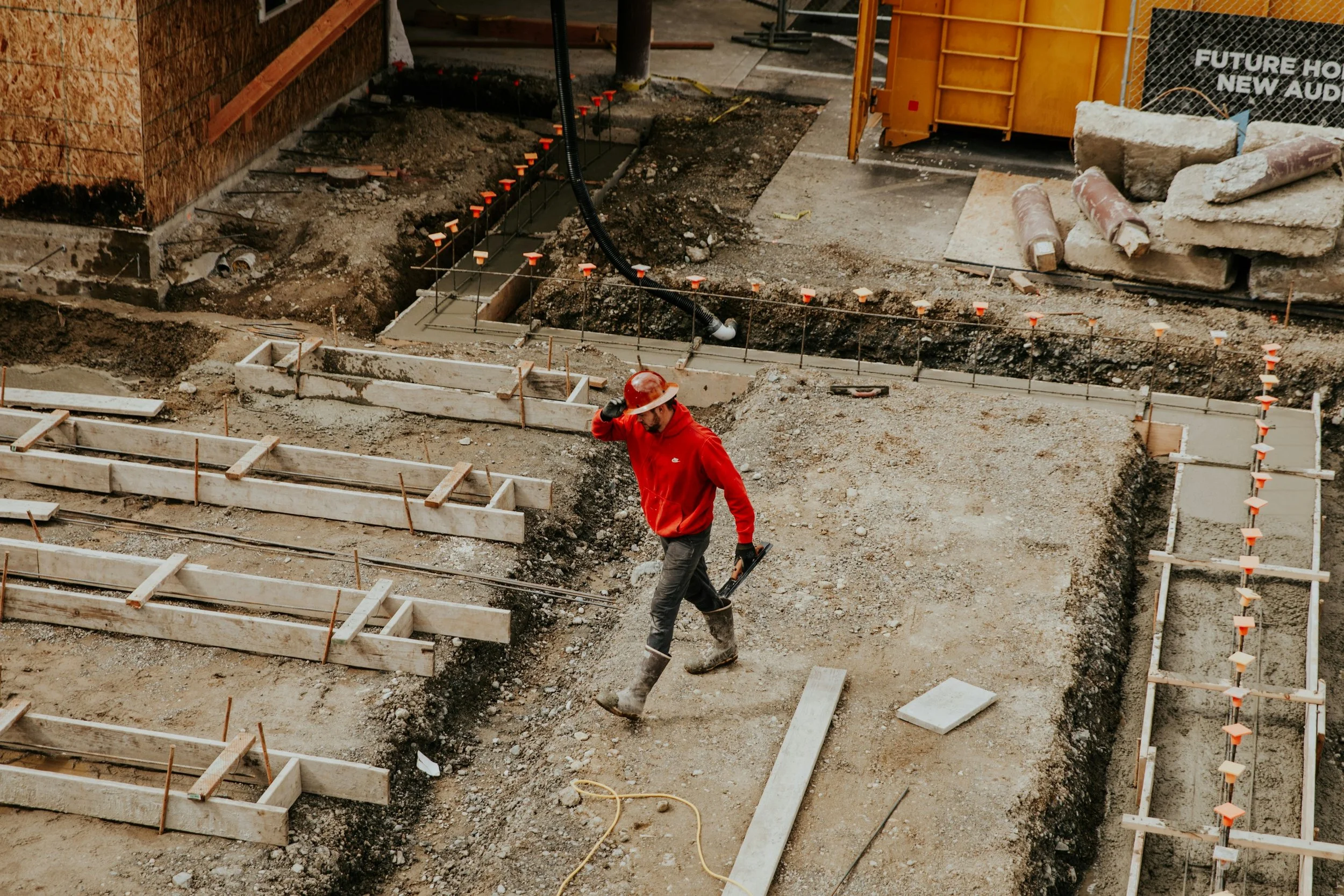 Construction site with worker in red jacket wearing a hard hat, working on concrete foundation with wooden formwork and safety stakes, surrounded by dirt and construction materials.