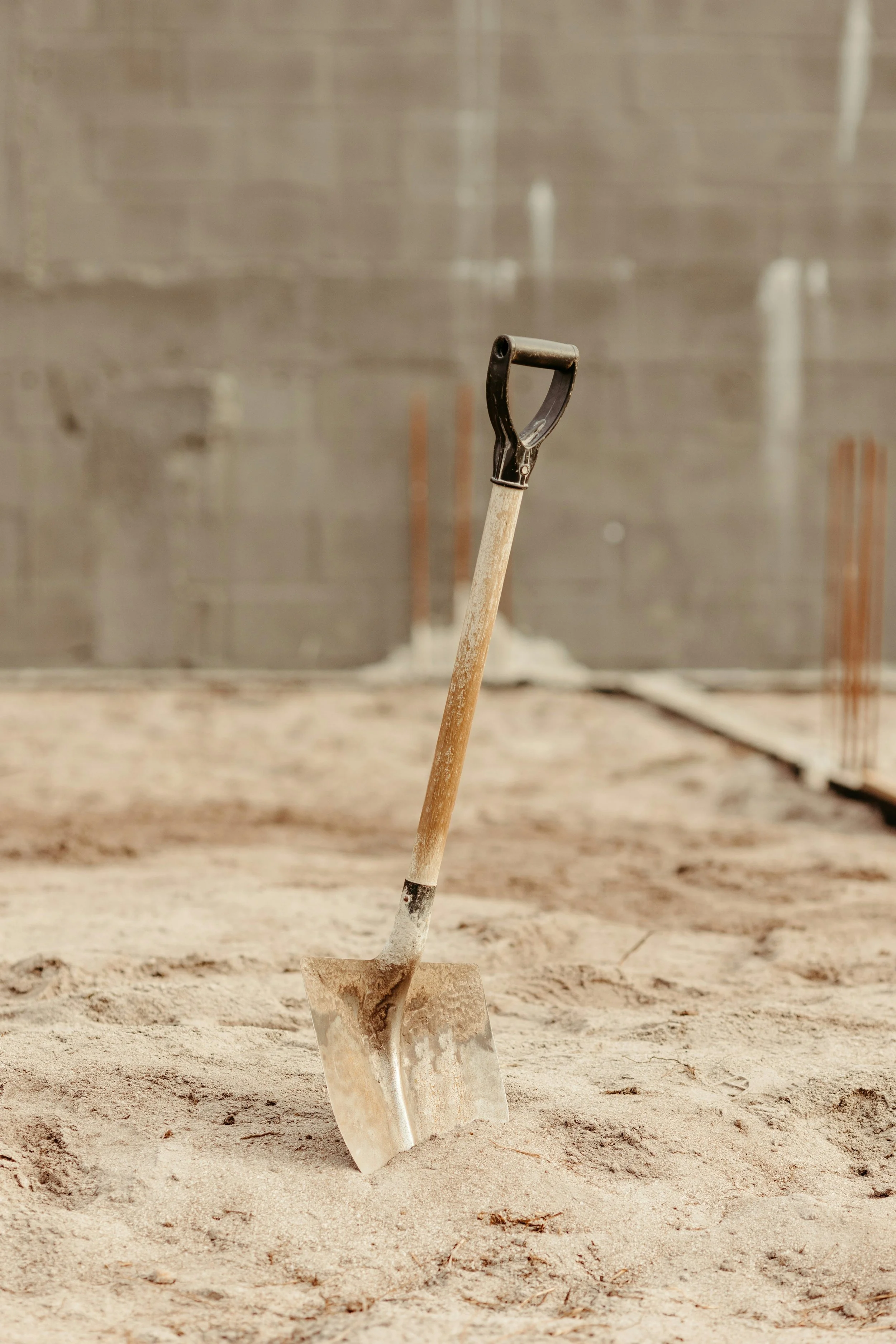 A shovel stands upright in dry, light-colored dirt at a construction site, with a gray concrete wall in the background.