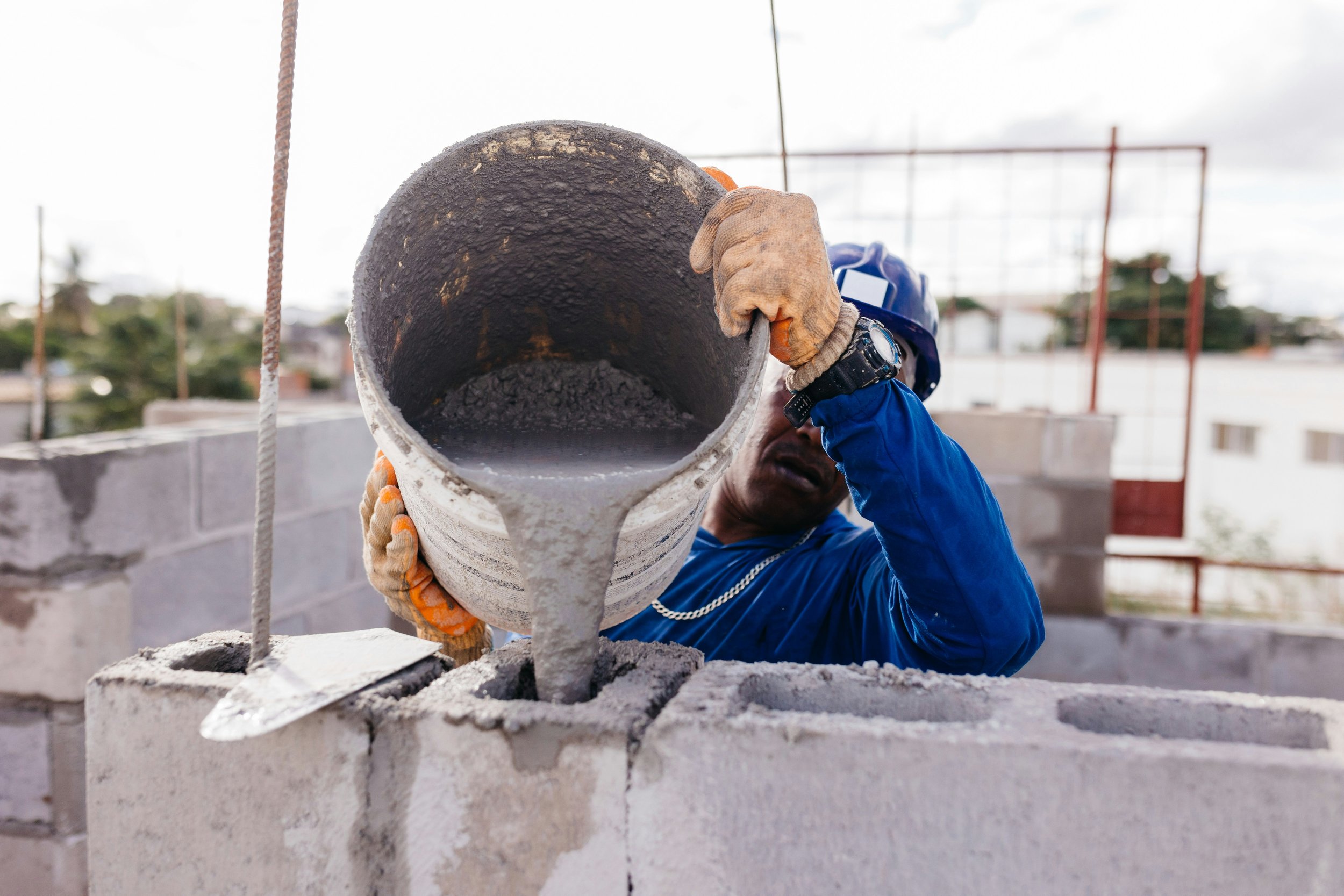 A construction worker pouring concrete from a bucket on a building site with a cloudy sky and scaffolding in the background.