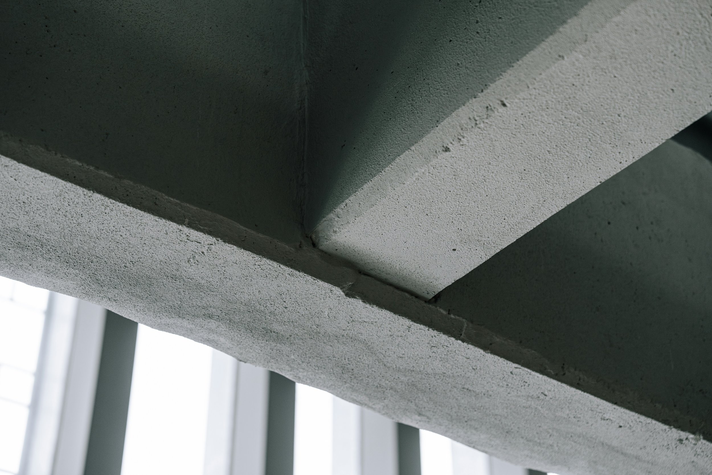 Close-up of the corner of a concrete ceiling with textured surface and visible joints, with part of a white vertical support structure in the background.