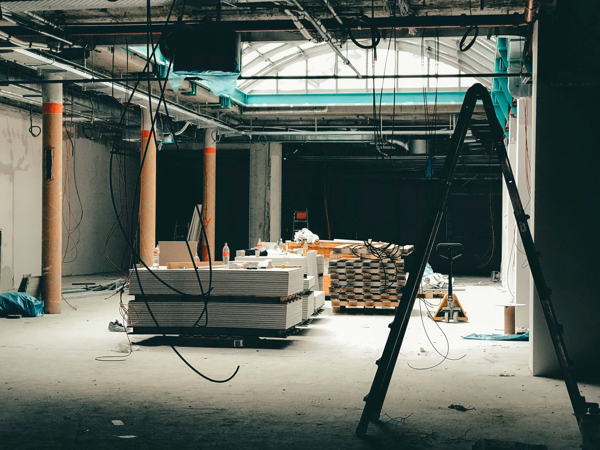 Interior of a building under construction with exposed ceiling pipes, wiring, and construction materials on the floor.