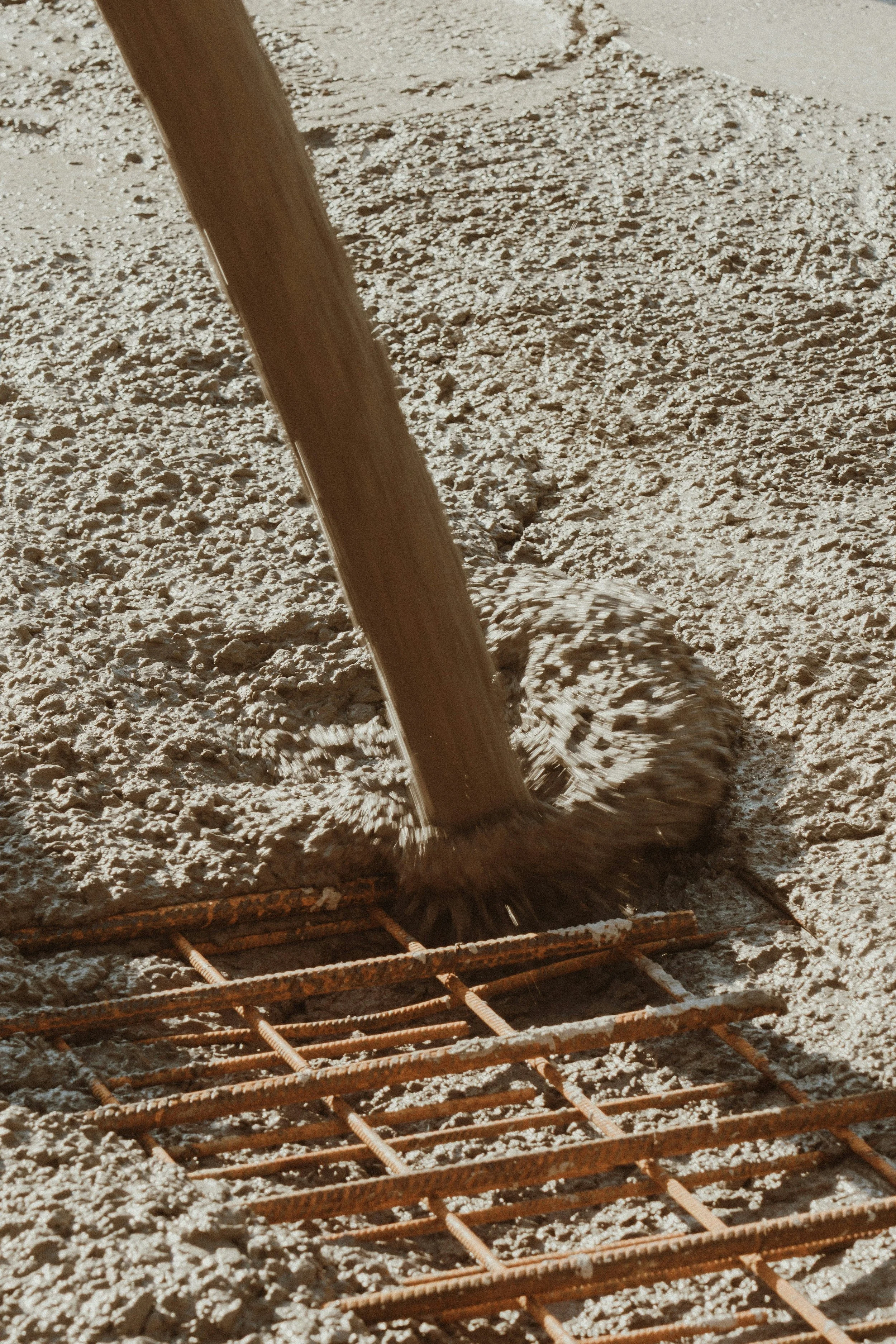 A construction site showing a steel reinforcement grid, a wheelbarrow, and a shovel in a sandy area, with a wooden-handled tool being used to move or spread gravel or dirt.