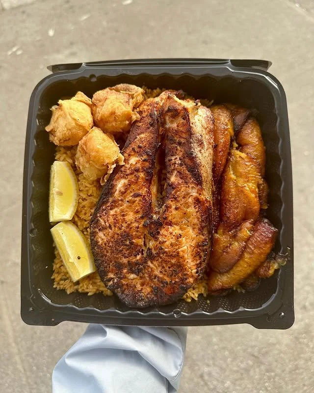 Fried fish fillet with two lemon wedges, fried chicken pieces, rice, and fried plantains in a black container.