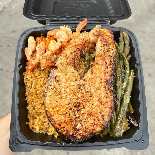 Black takeout container with grilled salmon filet, shrimp, rice, and green beans.