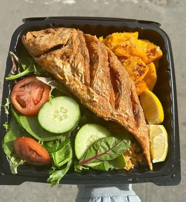 Fried fish fillet with side salad, cherry tomatoes, cucumber slices, greens, roasted plantains, and lemon wedges in a black takeout box.
