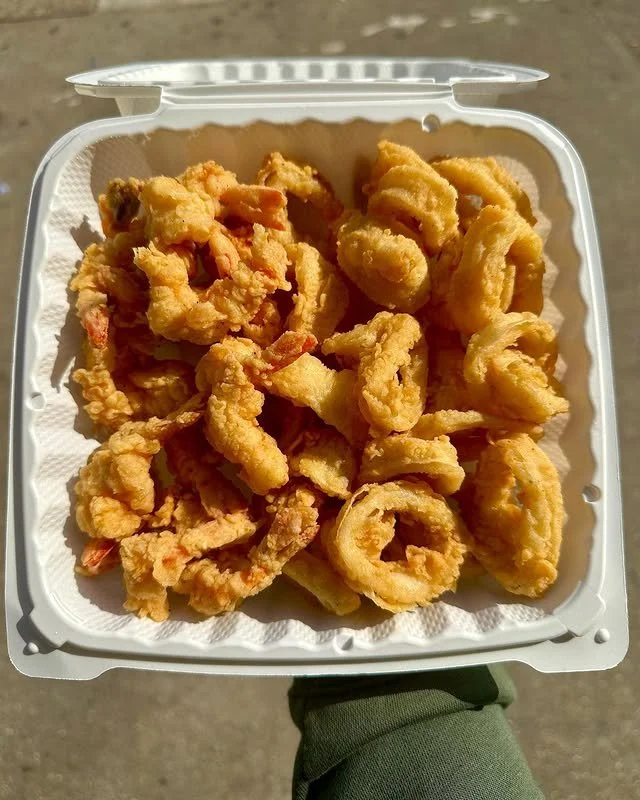 A white takeout container filled with fried calamari rings and pieces, placed outdoors.