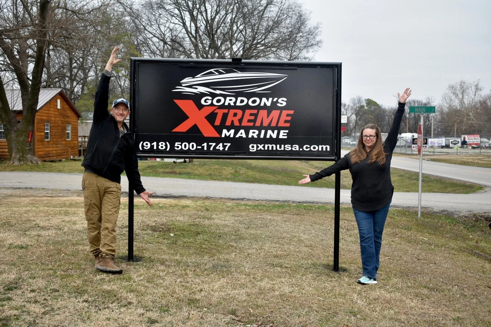 Two people, a man and a woman, posing next to a large sign for Gordon's Xtreme Marine, with the man pointing upwards and the woman extending her arms towards the sign, in an outdoor area with trees, grass, and a road in the background.