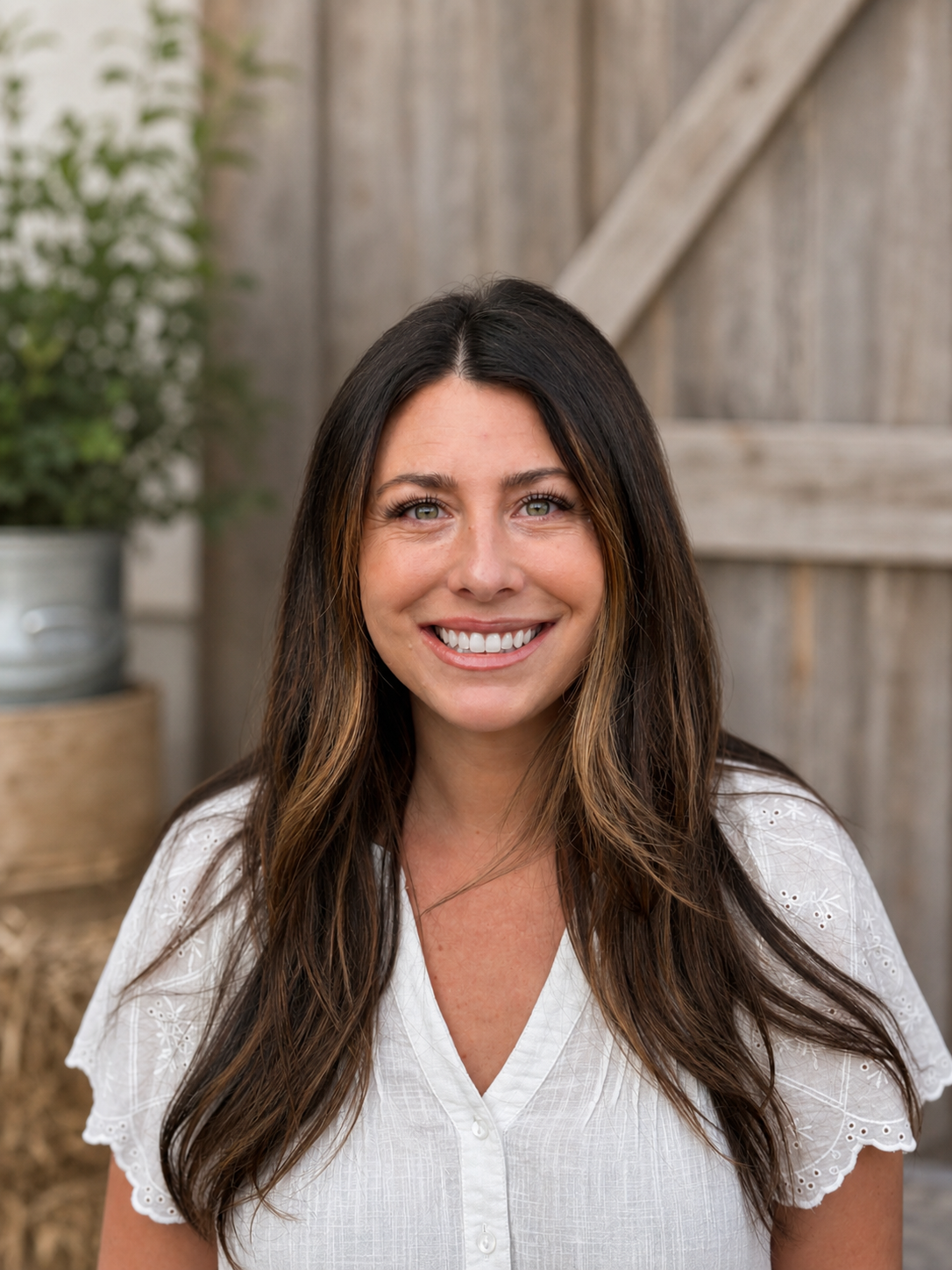 A woman with long brown hair and green eyes smiling, wearing a white blouse, standing outdoors near a wooden fence and potted plant in a rustic setting.