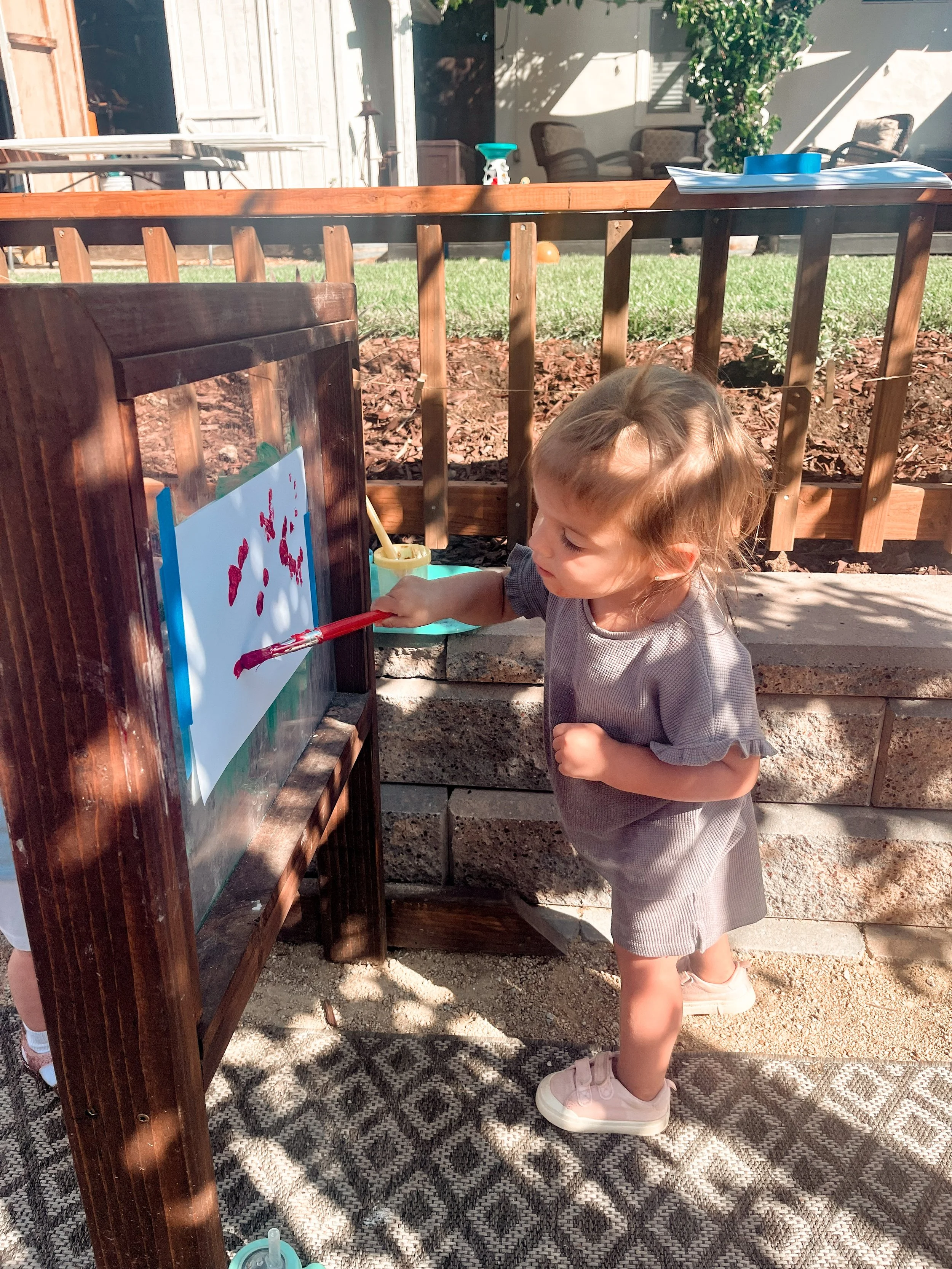 A young girl painting in an outdoor area with sunlight, a wooden fence, and patio furniture in the background. She is wearing a gray dress and pink sneakers.