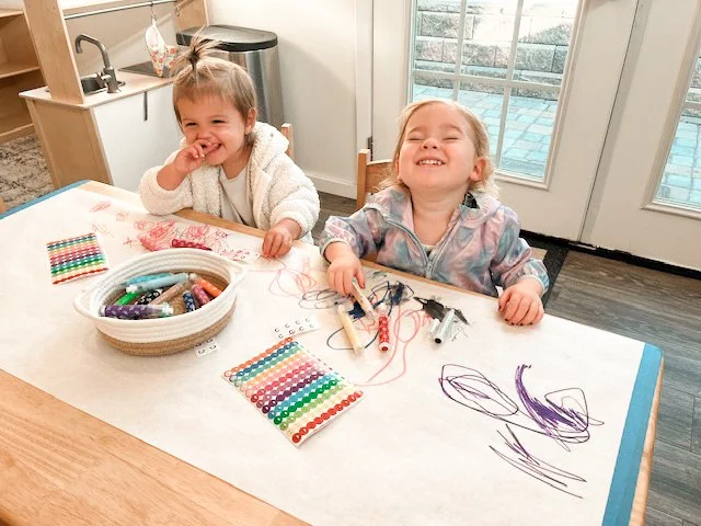 Two young girls sitting at a table, smiling and drawing with colorful crayons on paper, with a basket of crayons and some beaded bracelets on the table, in a bright room with a glass door leading outside.
