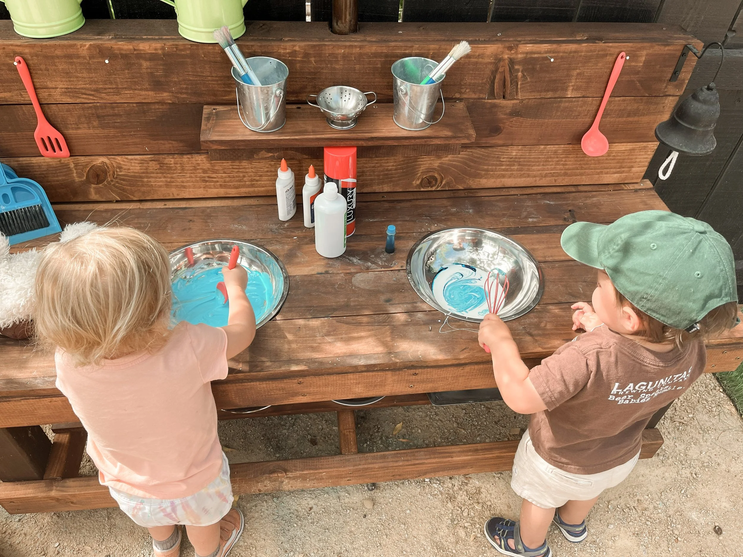 Two children are standing at a wooden table, stirring blue slime in metal bowls with red spatulas. The table has bottles of glue, a small container, and brushes. The children are wearing casual clothing and hats, and the setting appears to be outdoor