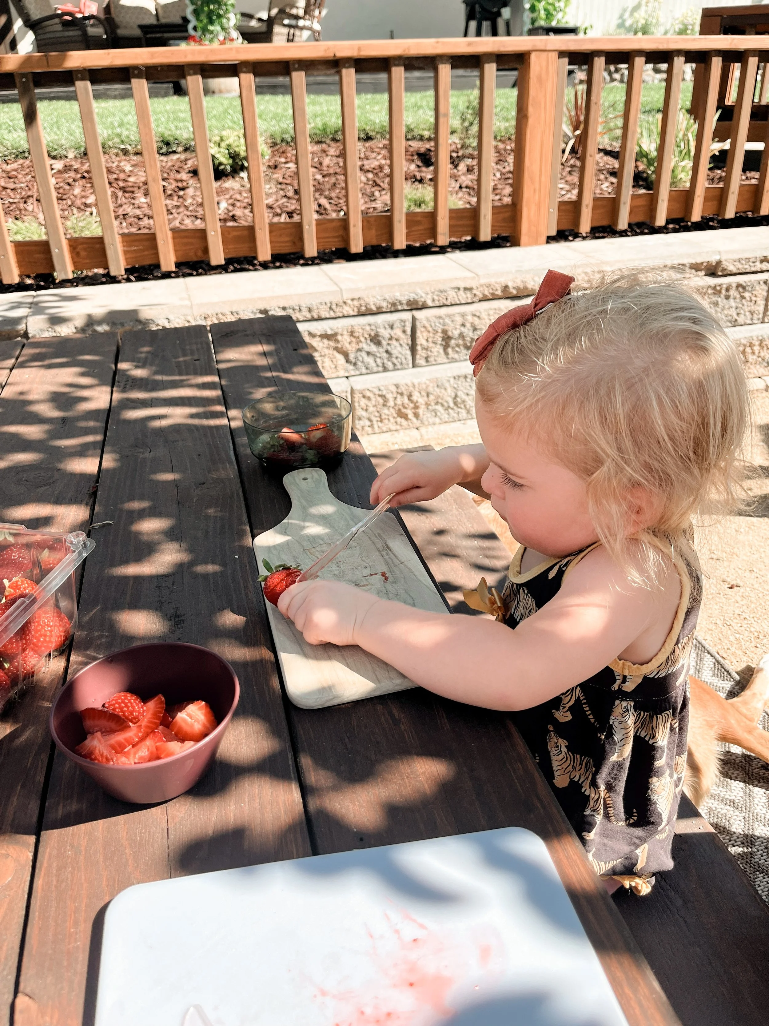 A young girl with blonde hair, wearing a black and beige dress with a bow, is cutting strawberries on a white cutting board outdoors. There are bowls of sliced strawberries on a dark wooden table, and a bowl of whole strawberries nearby. A fence and 