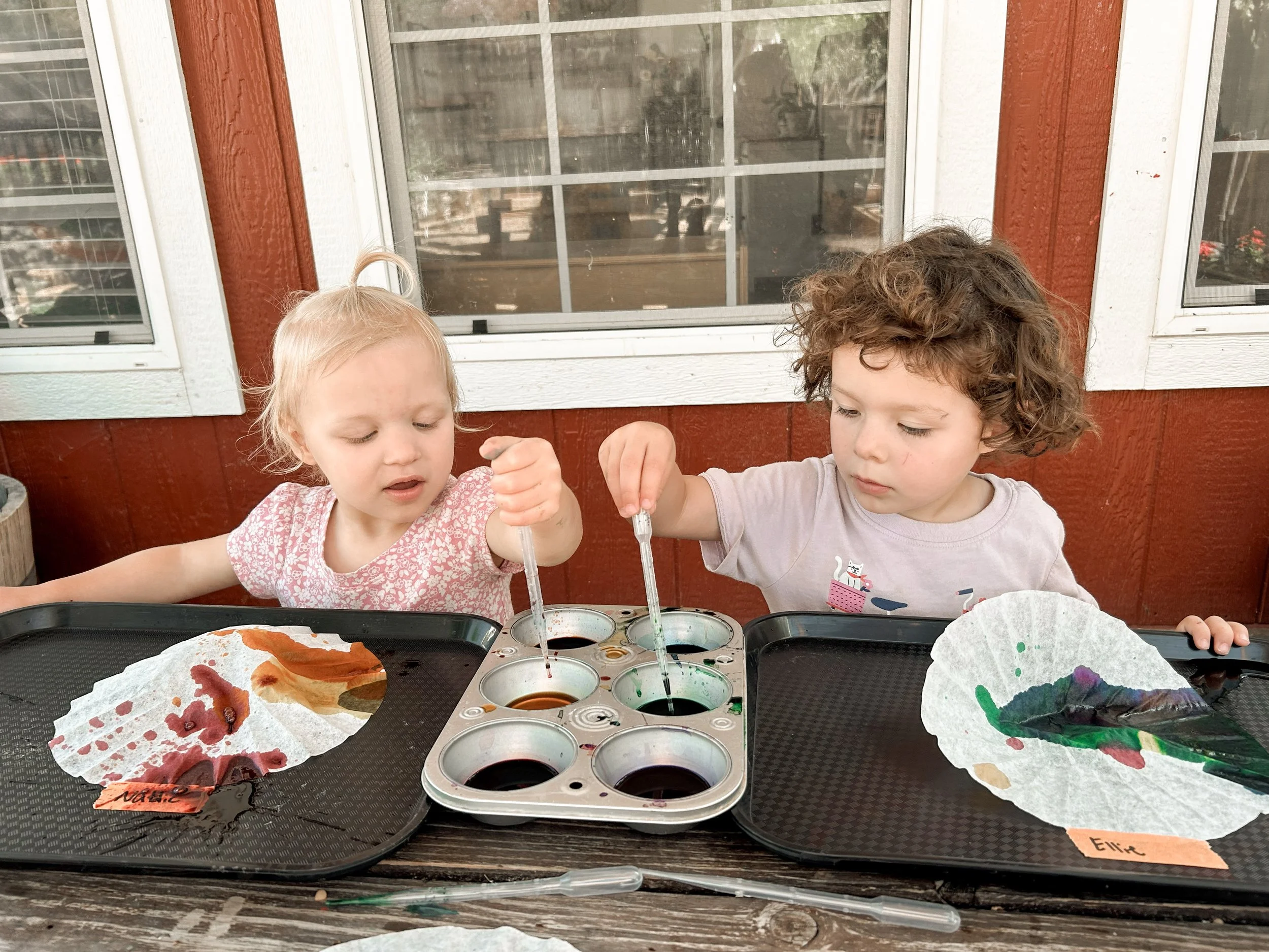 Two young children are sitting at a table outside, mixing watercolors in a palette. The girl on the left, with blonde hair, is wearing a pink floral top, and the boy on the right, with curly brown hair, is wearing a light gray shirt with a graphic pr