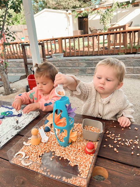 Two young children playing with a bean sensory bin outdoors at a wooden table.