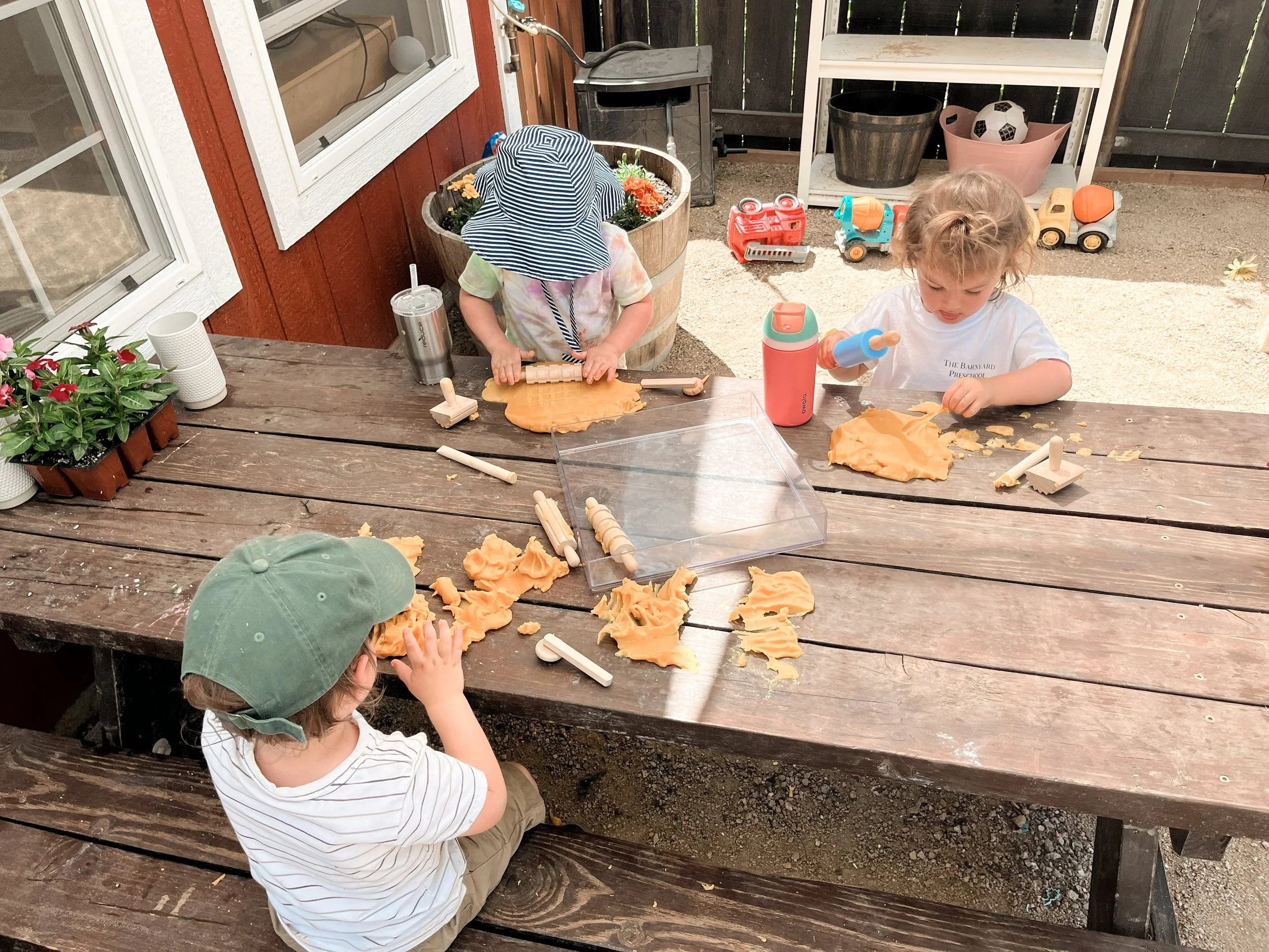 Two young children playing with orange-colored dough at a wooden table outdoors. One child is rolling dough with a wooden rolling pin, wearing a large sun hat, while the other is sitting and looking at the dough, wearing a striped shirt and a green c