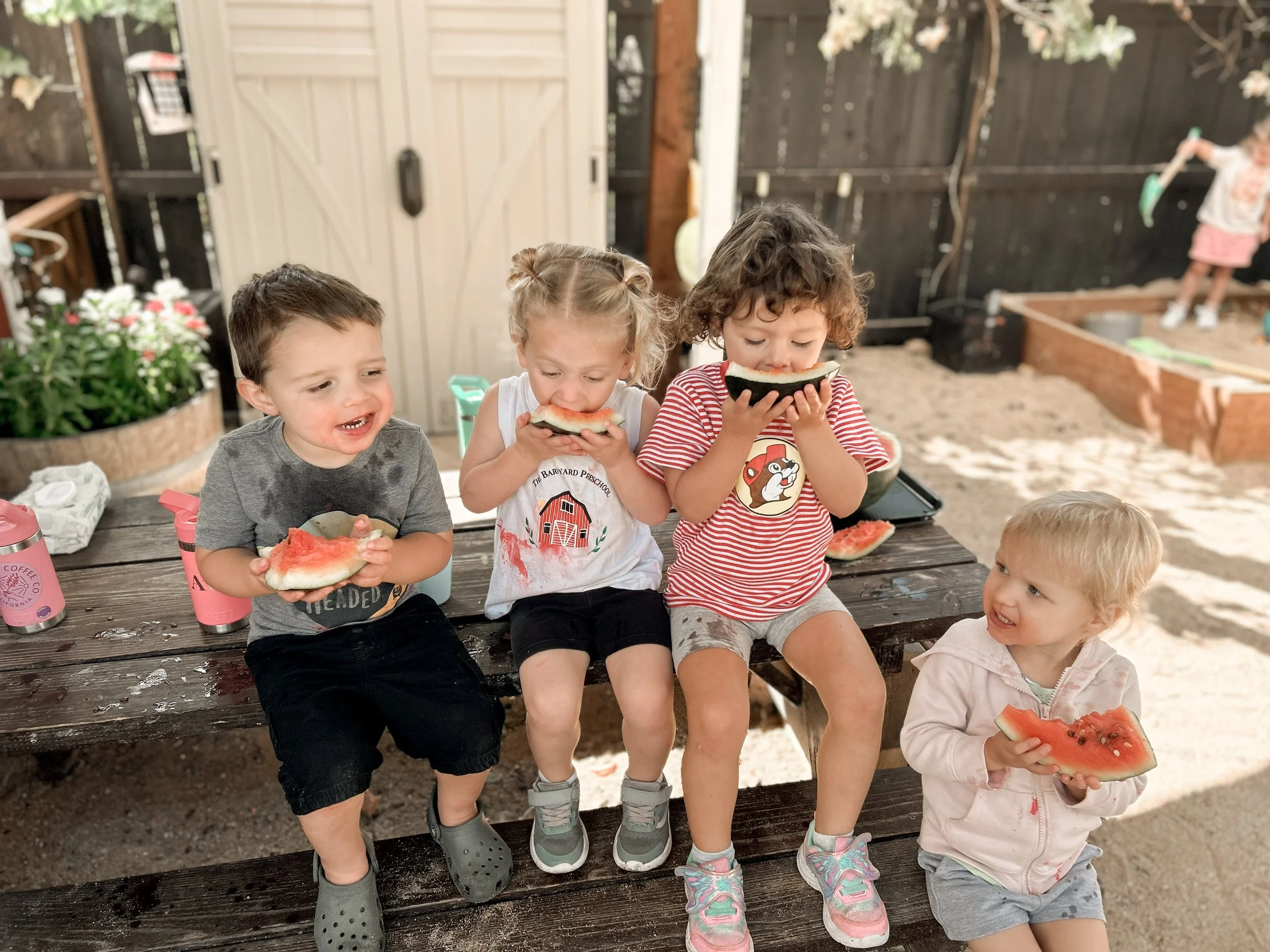 Four young children sit on a bench, eating slices of watermelon, outdoors in a backyard or garden area with a sandbox and wooden fences in the background.