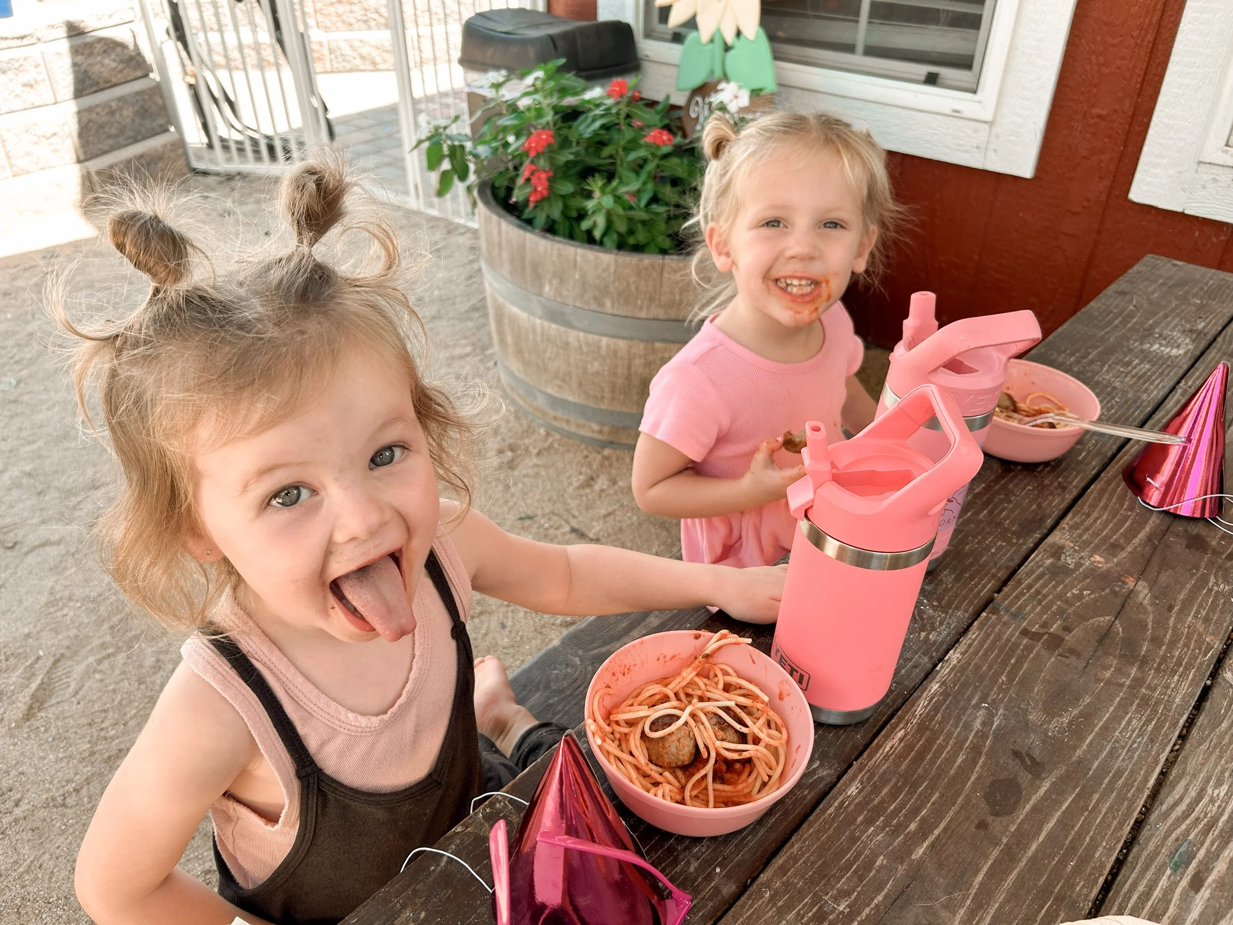 Two young girls enjoying a meal of spaghetti and meatballs at an outdoor table. One girl is smiling at the camera with food around her mouth, and the other girl is sticking her tongue out. There are pink cups, a pink water bottle, party hats, and a b