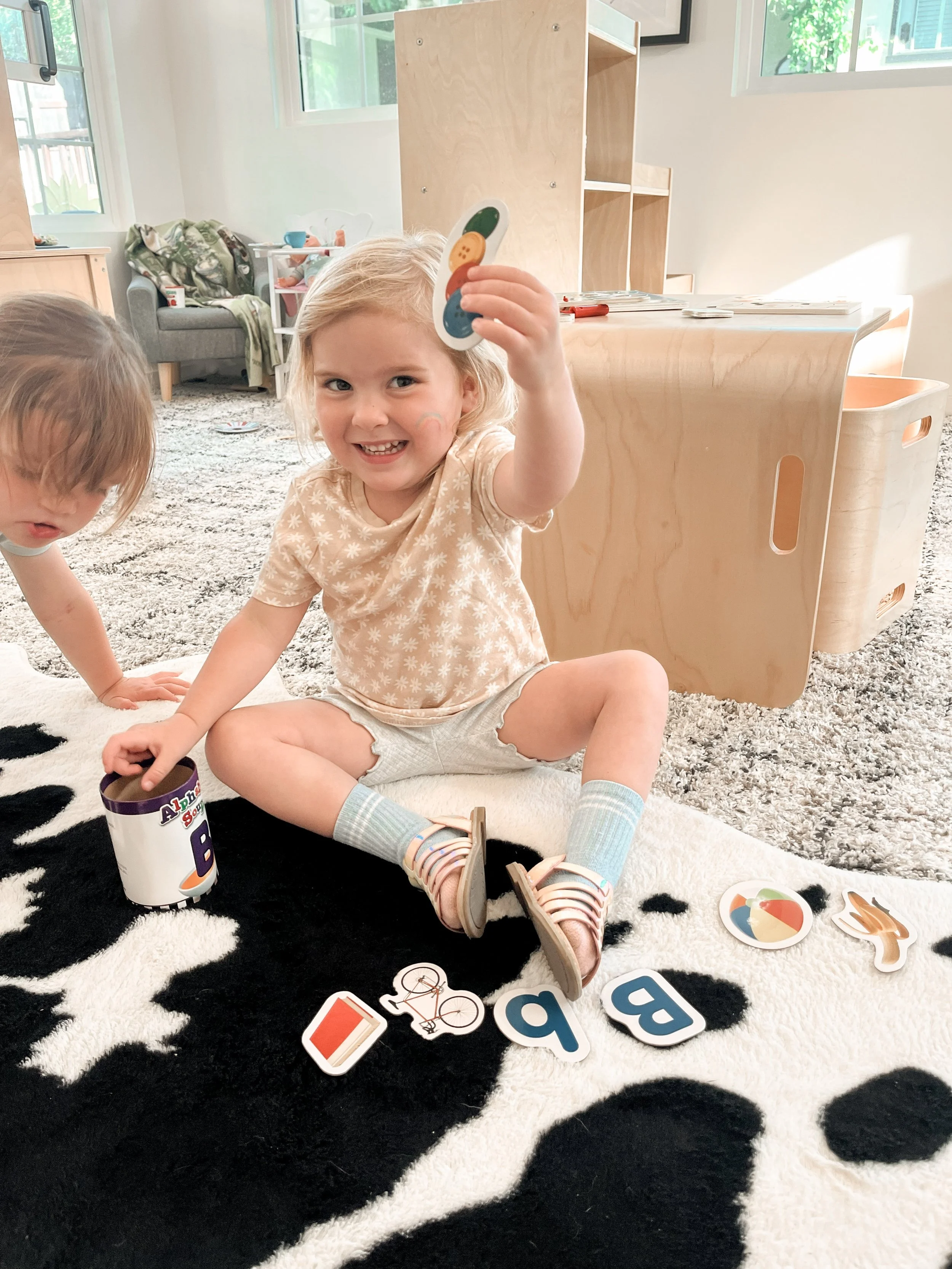 Two young children playing on a black and white rug with alphabet and picture flashcards inside a bright living room with large windows, wooden furniture, and a beige carpet in the background.