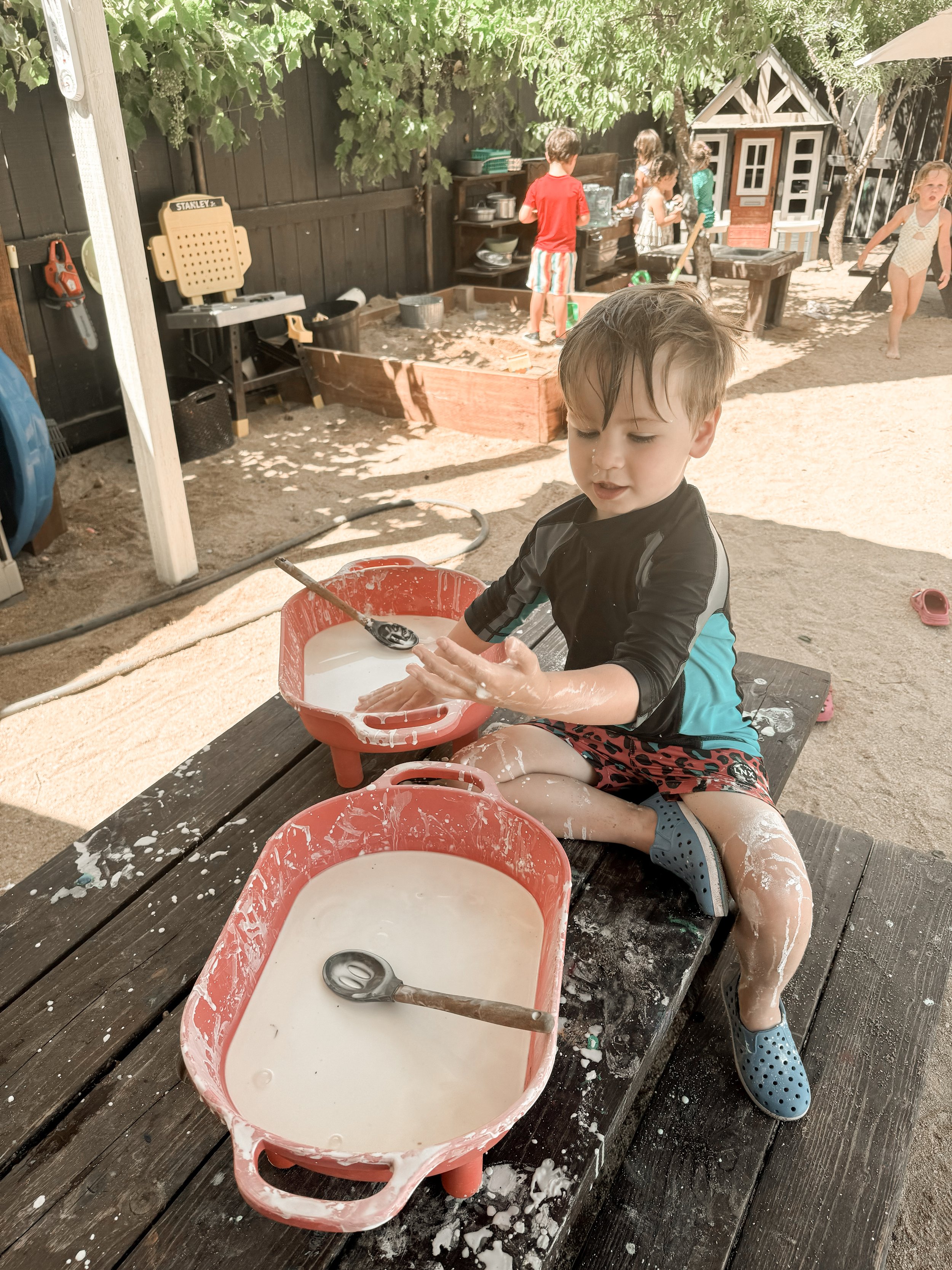 A young boy sitting on a wooden deck, engaging in a sensory activity with a red plastic tray filled with soapy water and a spoon inside. Behind him, there is a sandbox with children playing, a playhouse, and trees providing shade.