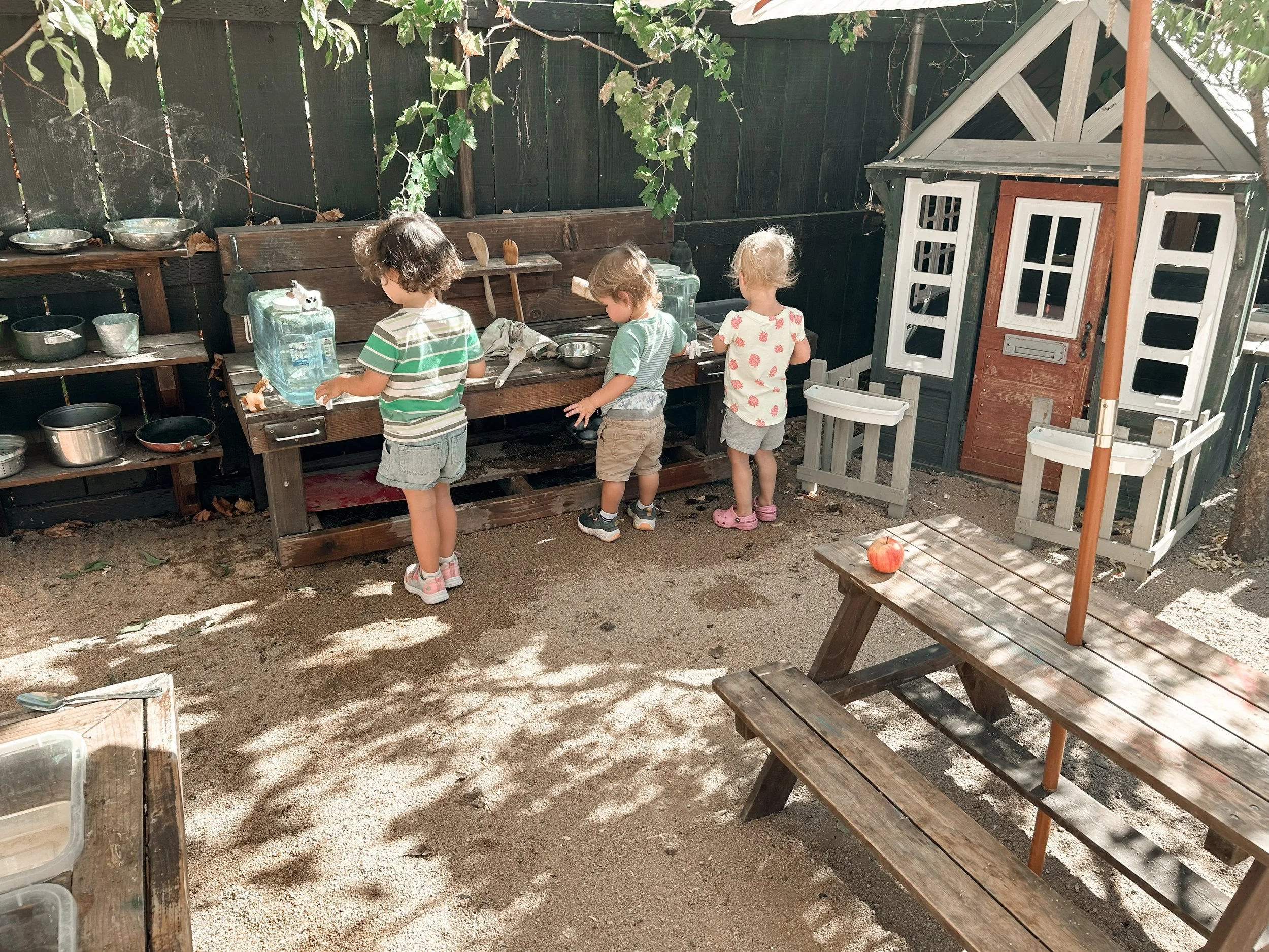 Four children playing outside near a wooden playhouse and sandbox on a sandy surface, surrounded by a wooden fence and shaded by a tree.