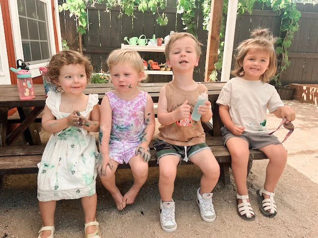 Four young children sitting on a wooden bench outdoors, smiling and holding small items. A backyard with a table, a shelf with kitchenware, and a fence with plants is visible in the background.