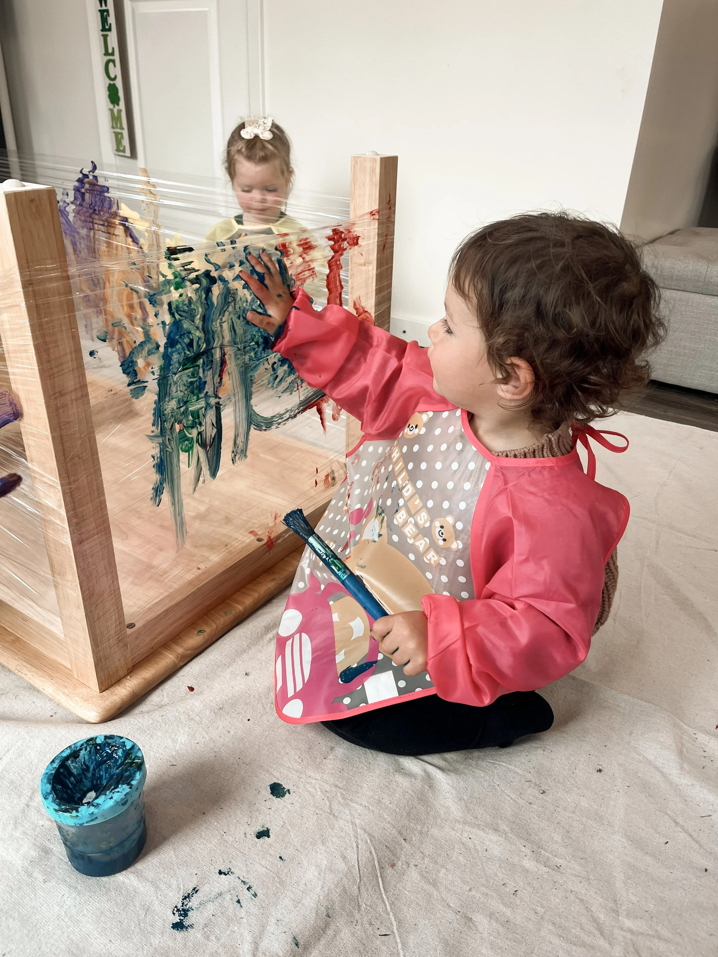 A young child kneeling on the carpeted floor wearing a pink apron with a brown bear design, painting on a wooden frame with a paintbrush, with another child behind the frame observing, and a blue paint cup nearby.