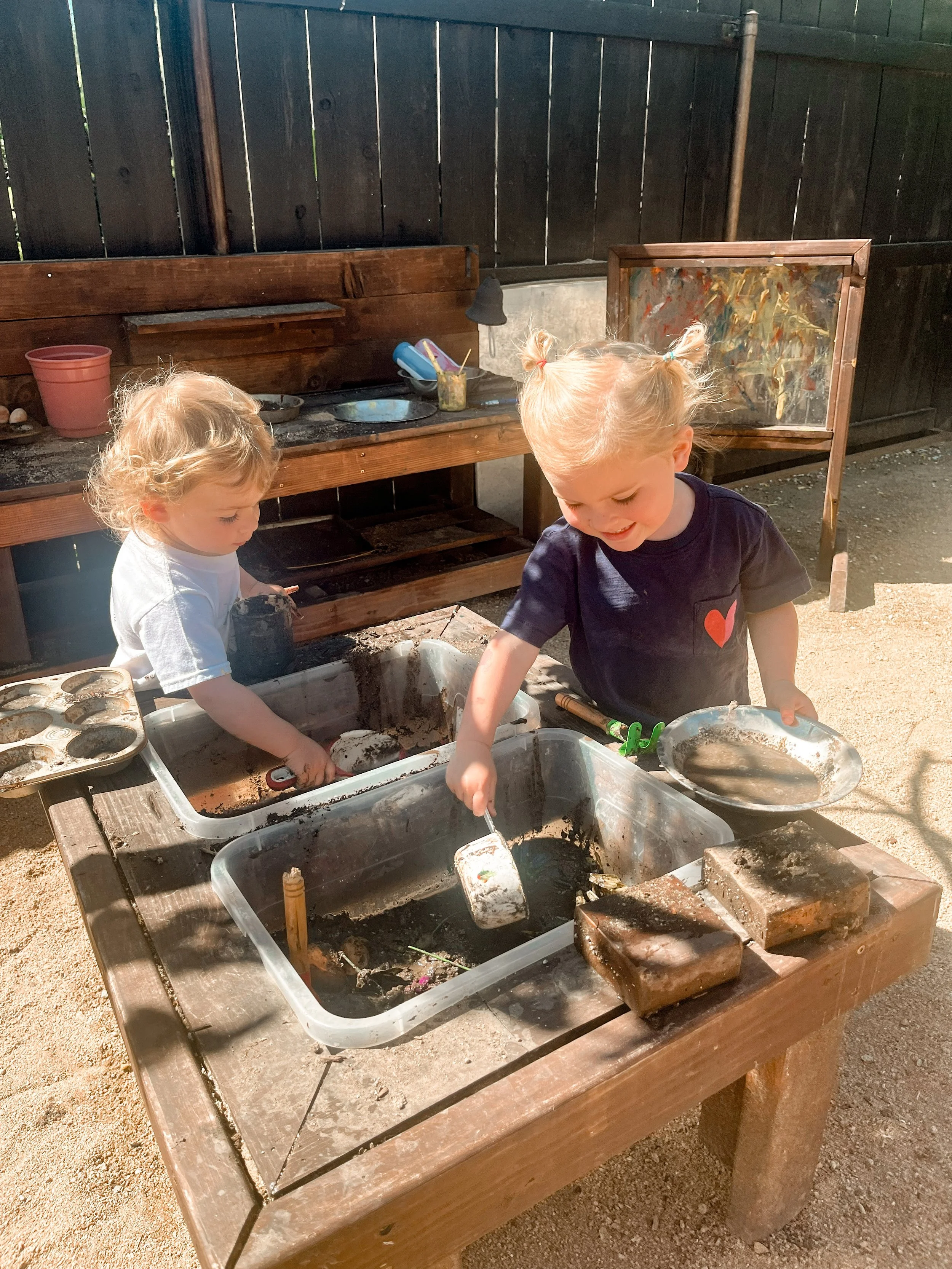 Two young children playing in an outdoor sandbox with toy shovels and pans, surrounded by soap and soil, with a wooden fence in the background.