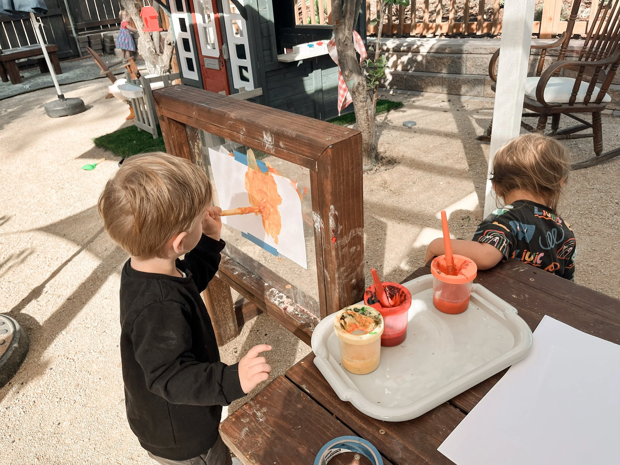 A young boy with blond hair painting on a piece of paper attached to a wooden easel outside, with cups of orange and red paint nearby. In the background, there is a girl in a dress, a small playhouse, chairs, a tree, and a sandbox area.