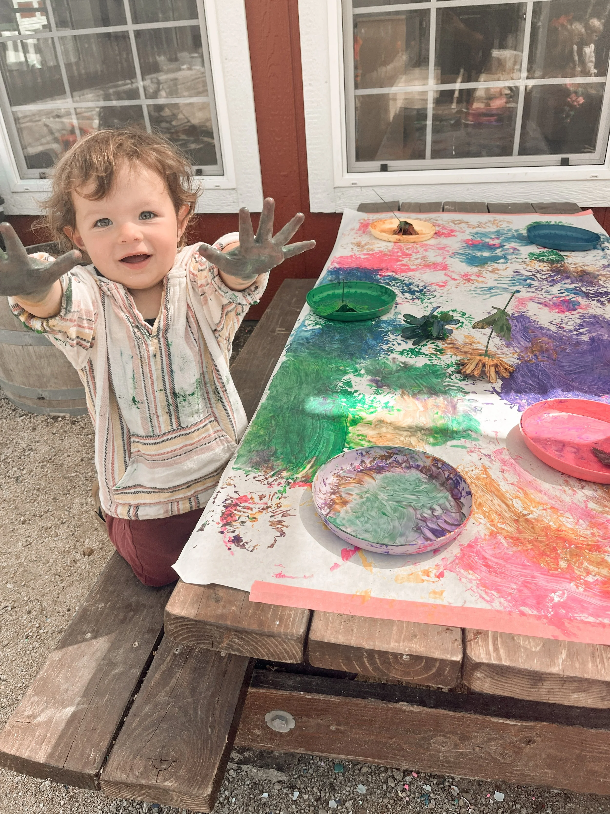 A young child with curly hair and a striped shirt looks happily at the camera with hands covered in green paint. The child is sitting at a wooden table covered with colorful, abstract finger-painting on paper, with bowls of different colored paints o