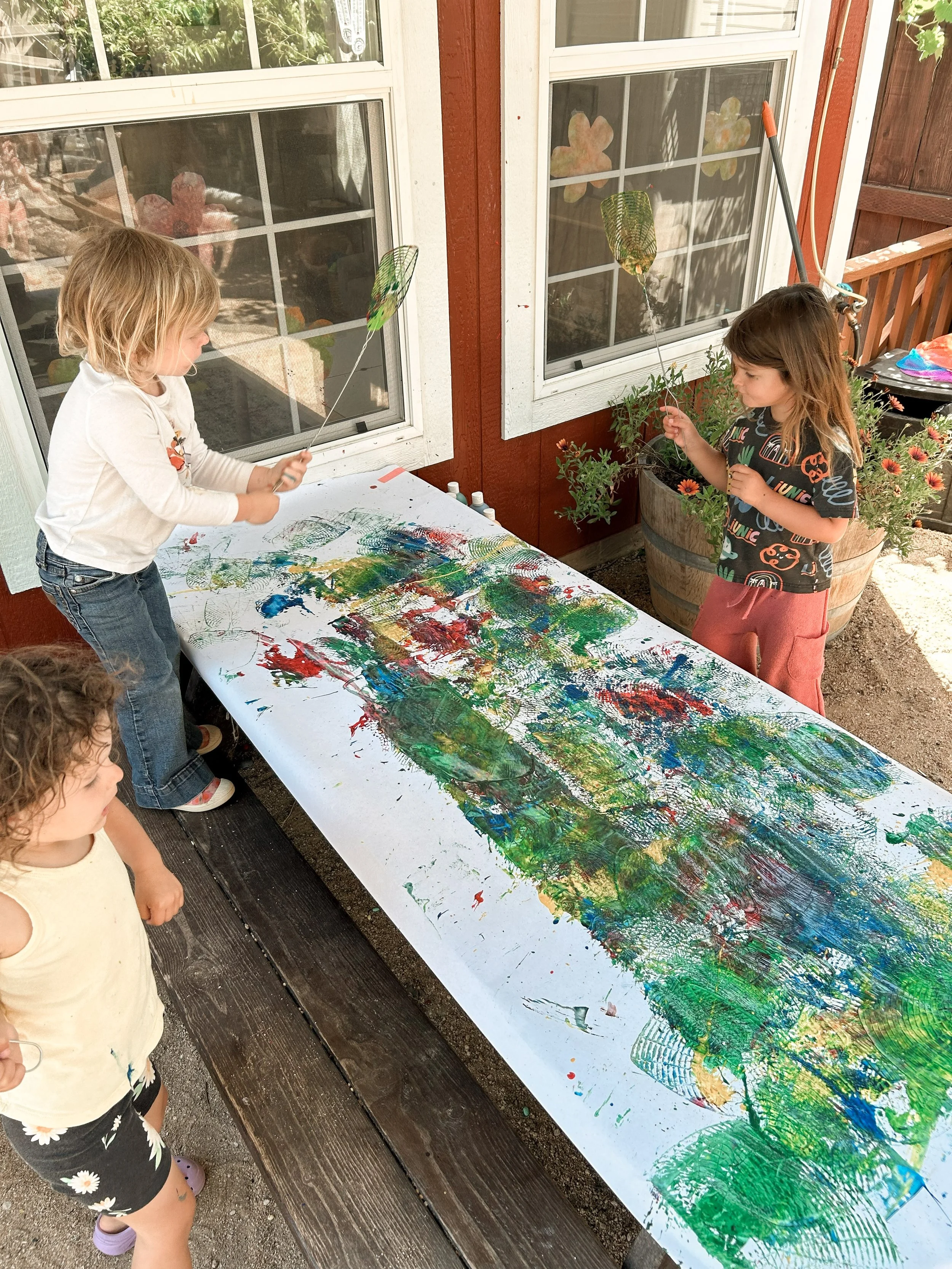 Two young girls are involved in a finger painting activity on a long sheet of paper outdoors, with paint and their fingers.