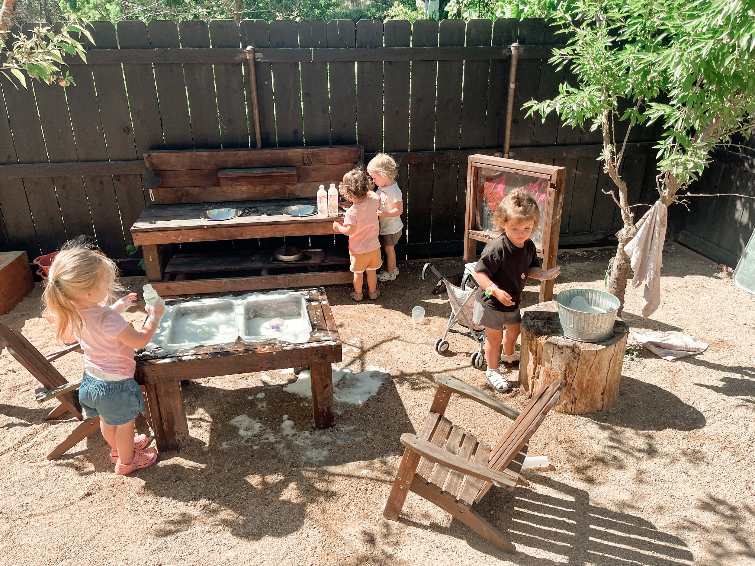 Children playing and engaging in outdoor activities in a backyard with a sandbox, a wooden bench, and a decorated wooden dragon.