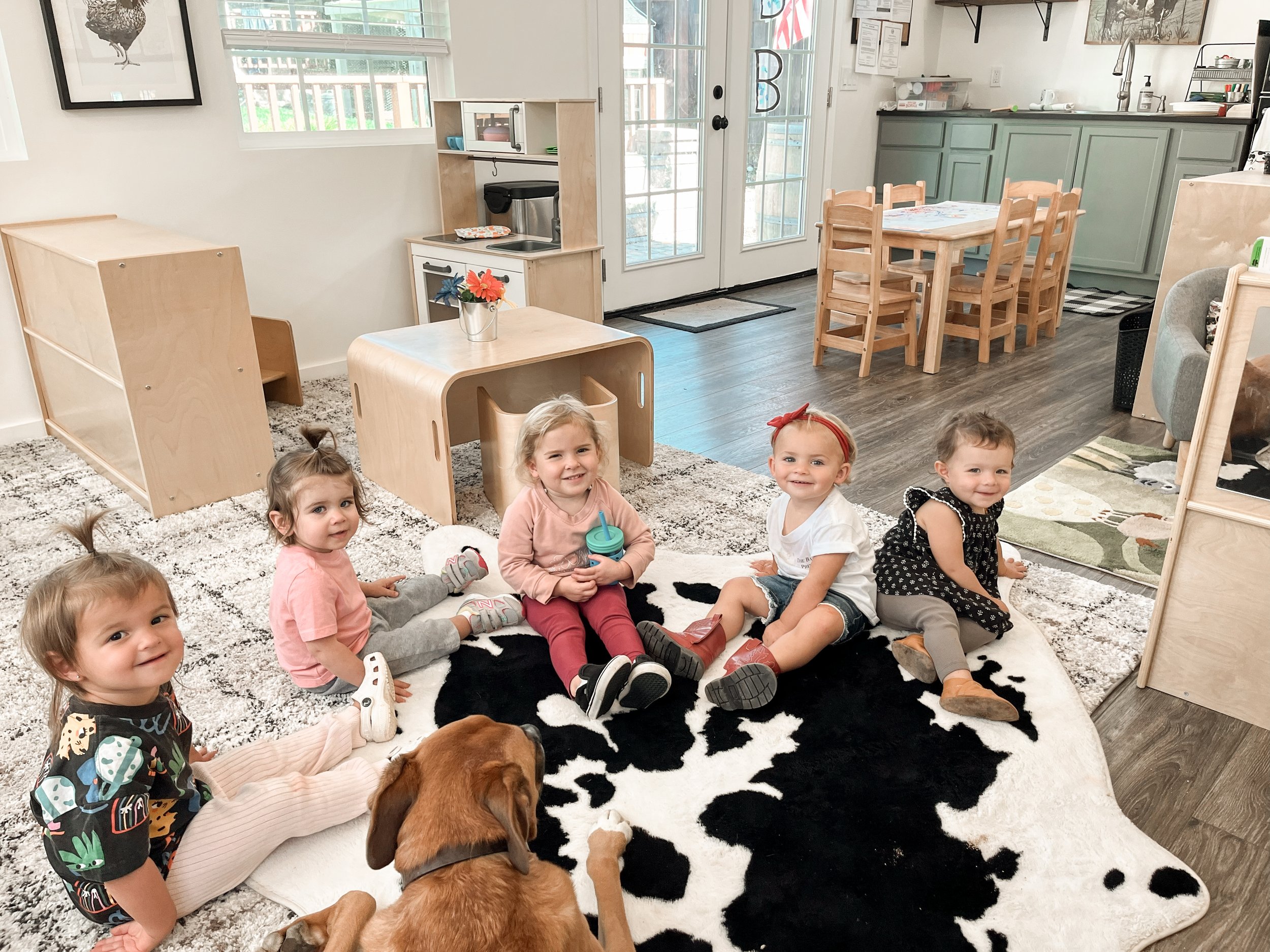 A group of young children sitting on a black and white cowhide rug in a playroom, smiling at the camera. There are several kids, some with light hair and one with red hair, and a brown dog lying down among them. The room has a wooden table, chairs, a