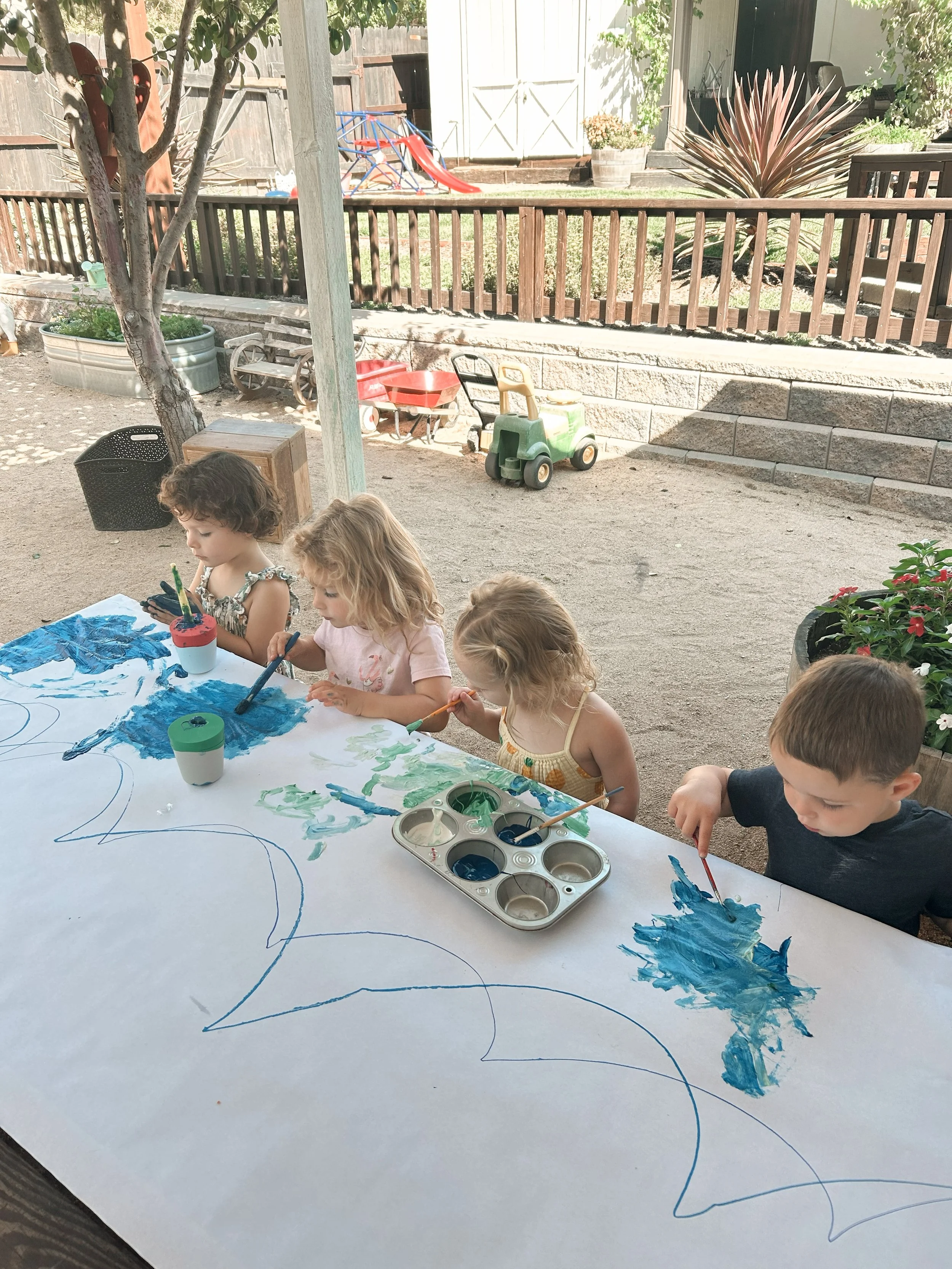 Children sitting at a table outdoors painting on a large sheet of paper with blue and green paint.