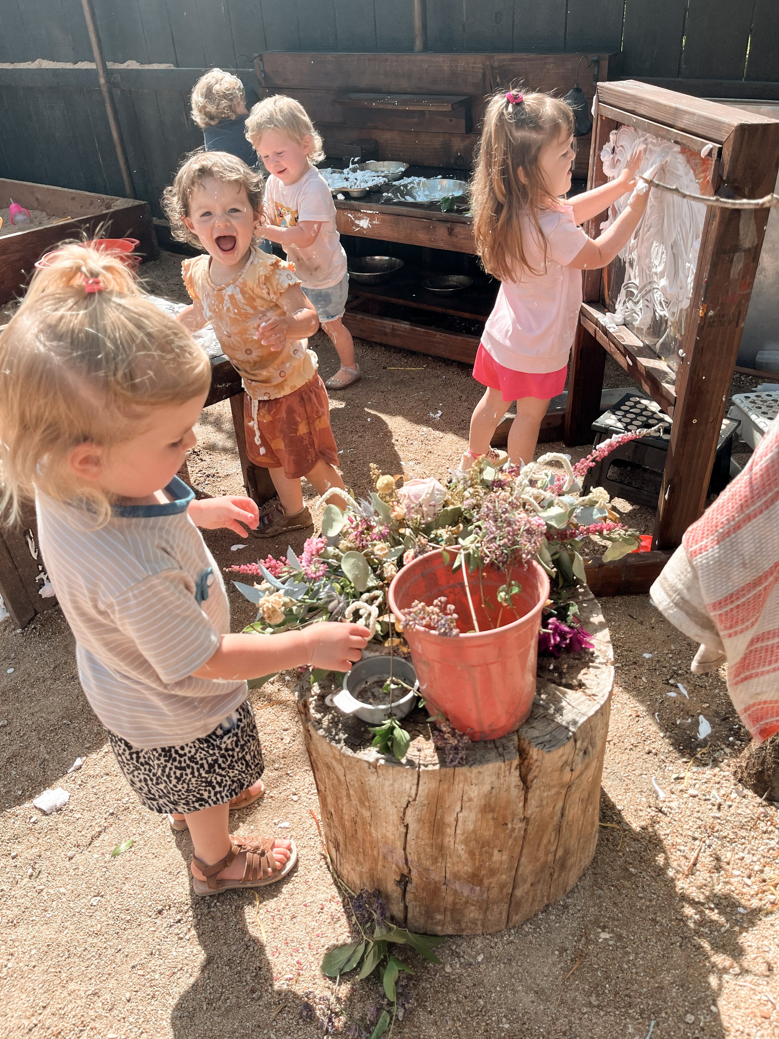 Children playing outdoors, engaging in a messy activity involving white foam or paint on a wooden frame and a tree stump with flowers in a pot, in a backyard or garden setting.